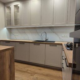 Manity Design Mateusz Łagiewa - Jasnoszara zabudowa kuchenna z drewnianym blatem, białym marmurowym panelem nad blatem i oświetleniem LED podszafkowym; widoczny zlew, bateria i płyta grzewcza.