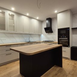 Manity Design Mateusz Łagiewa - Nowoczesna kuchnia z wyspą, jasne szafki z drewnianymi blatami, czarny sprzęt AGD i marmurowe ściany.