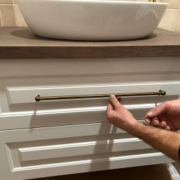 Manity Design Mateusz Łagiewa - Montaż uchwytu w złotej barwie do białej szafki łazienkowej pod umywalką nablatową z blatem w kolorze drewna.