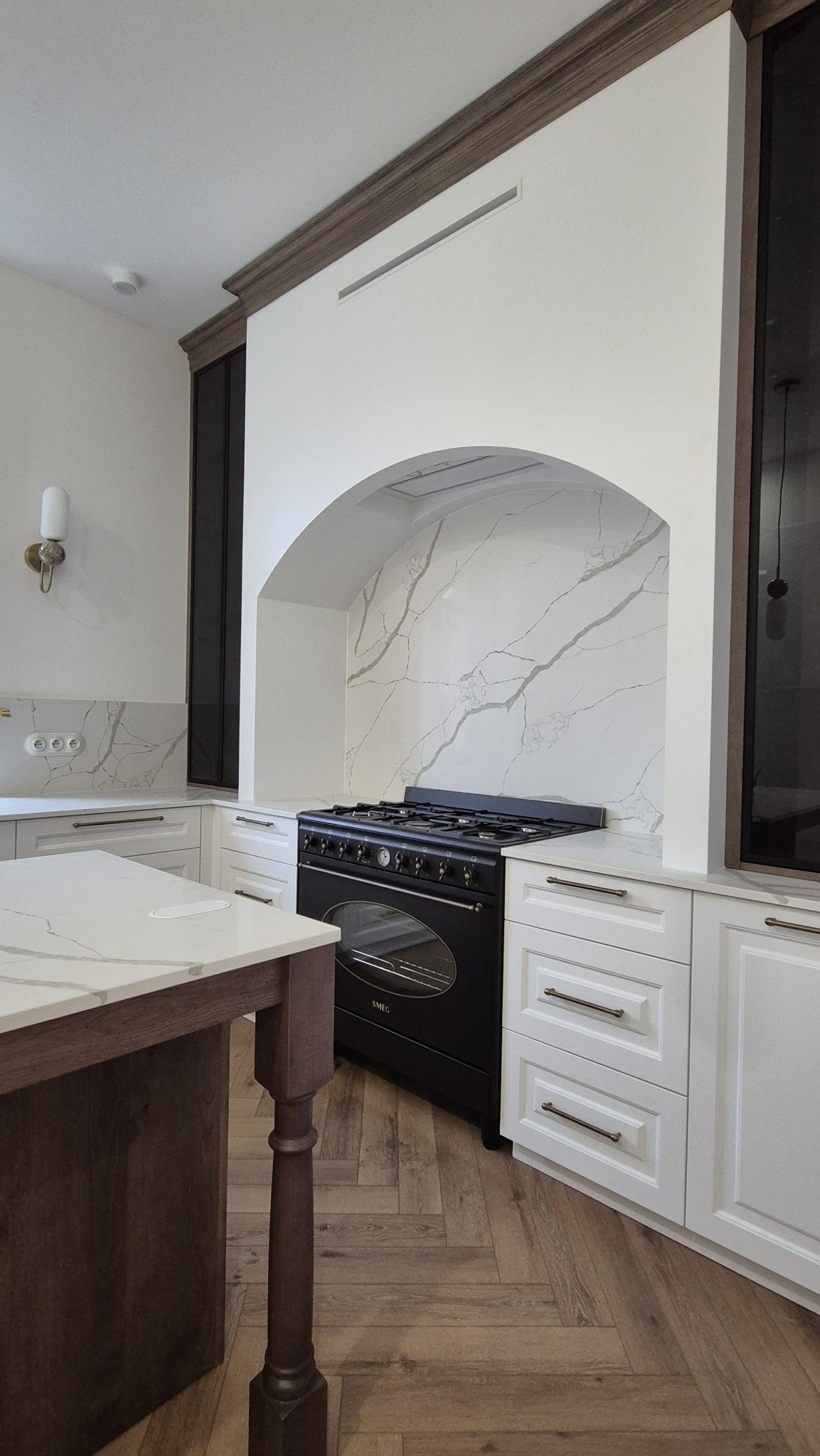 Elegancka kuchnia z czarnym piecem wolnostojącym SMEG w niszy z marmurowym tłem, białe szafki z poziomymi uchwytami i drewniany blat wyspy z marmurowym wykończeniem.