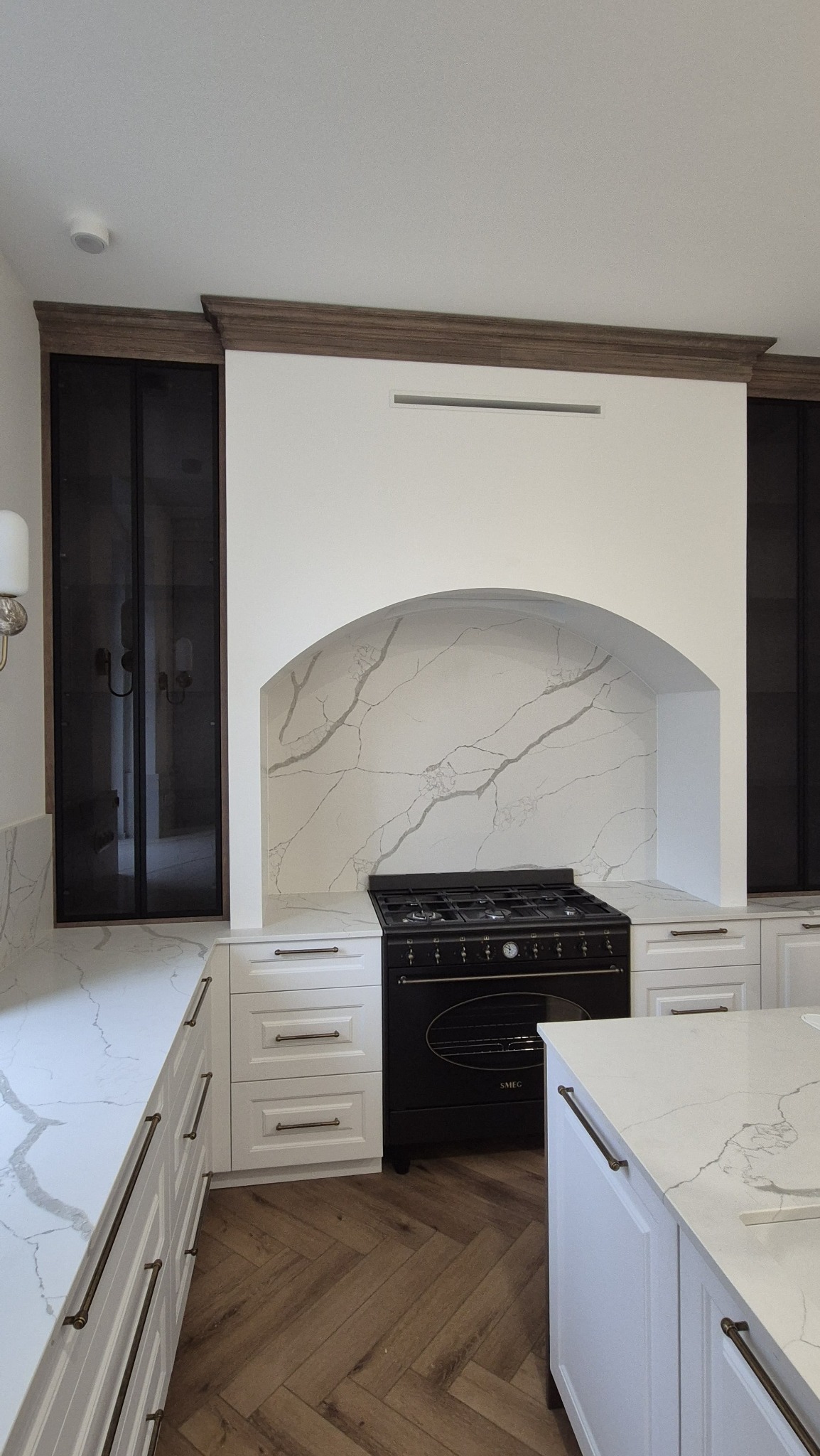 Elegancka kuchnia z białymi szafkami, blatem z marmuru i czarnym piecem SMEG, wykończona drewnianymi elementami dekoracyjnymi i podłogą w jodełkę.