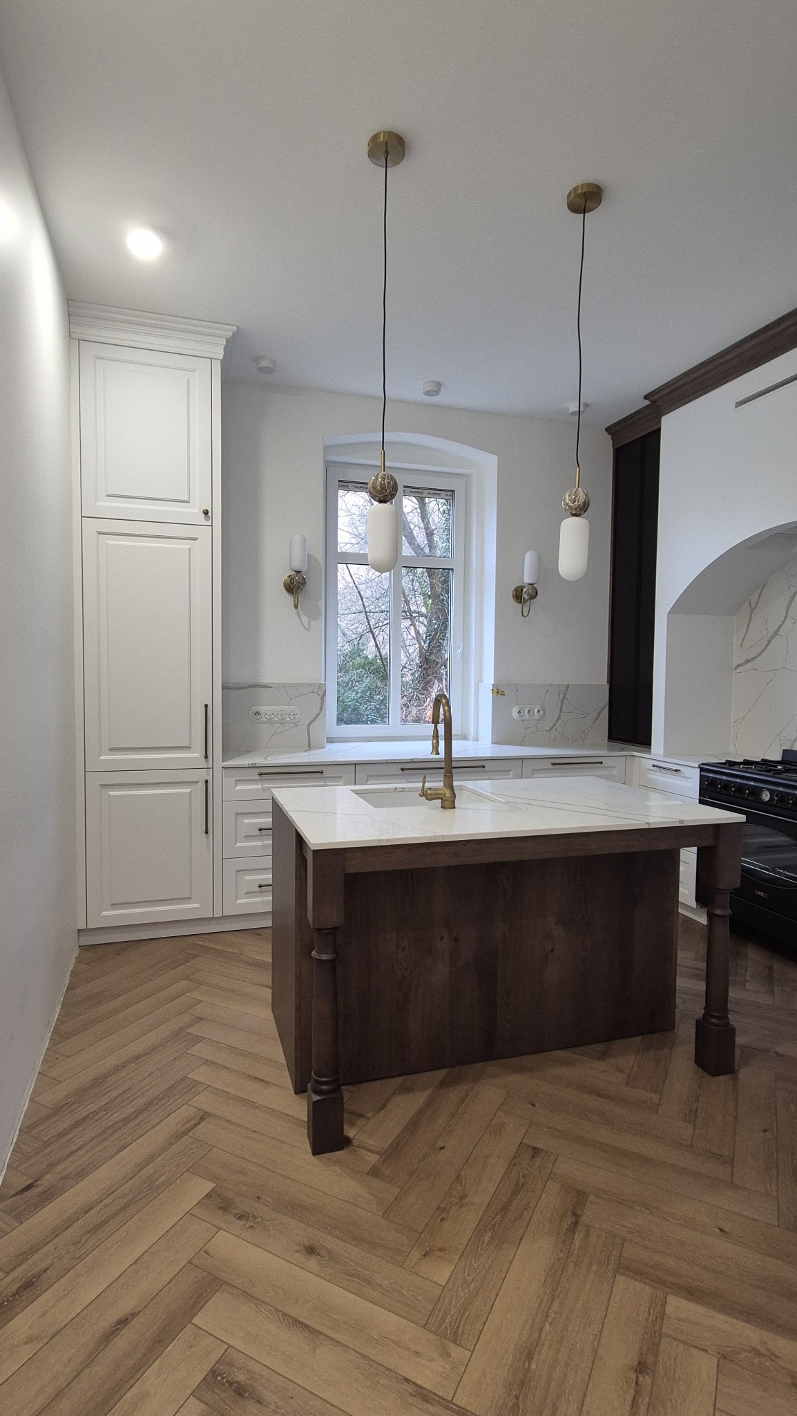 Nowoczesna kuchnia z wyspą, białymi szafkami do sufitu i parkietem w jodełkę; złote lampy wiszące i bateria; widok na okno.