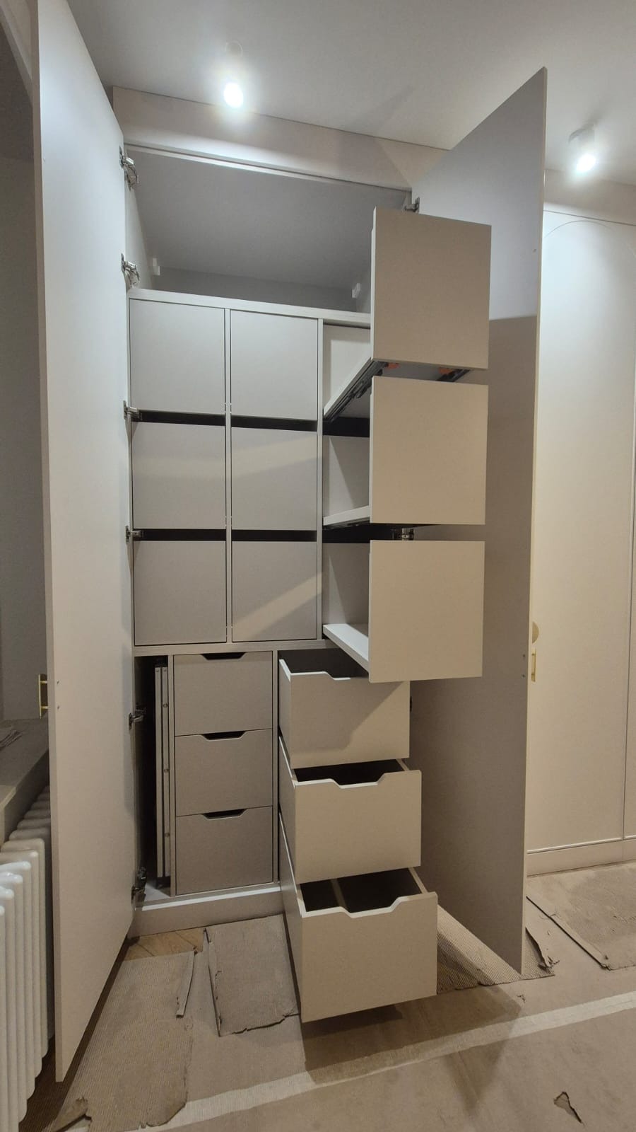 Wnętrze szafy wnękowej z wysuwanymi szufladami i półkami, widoczne mechanizmy wysuwu, jasne kolory, fragment grzejnika, podłoga w trakcie remontu.