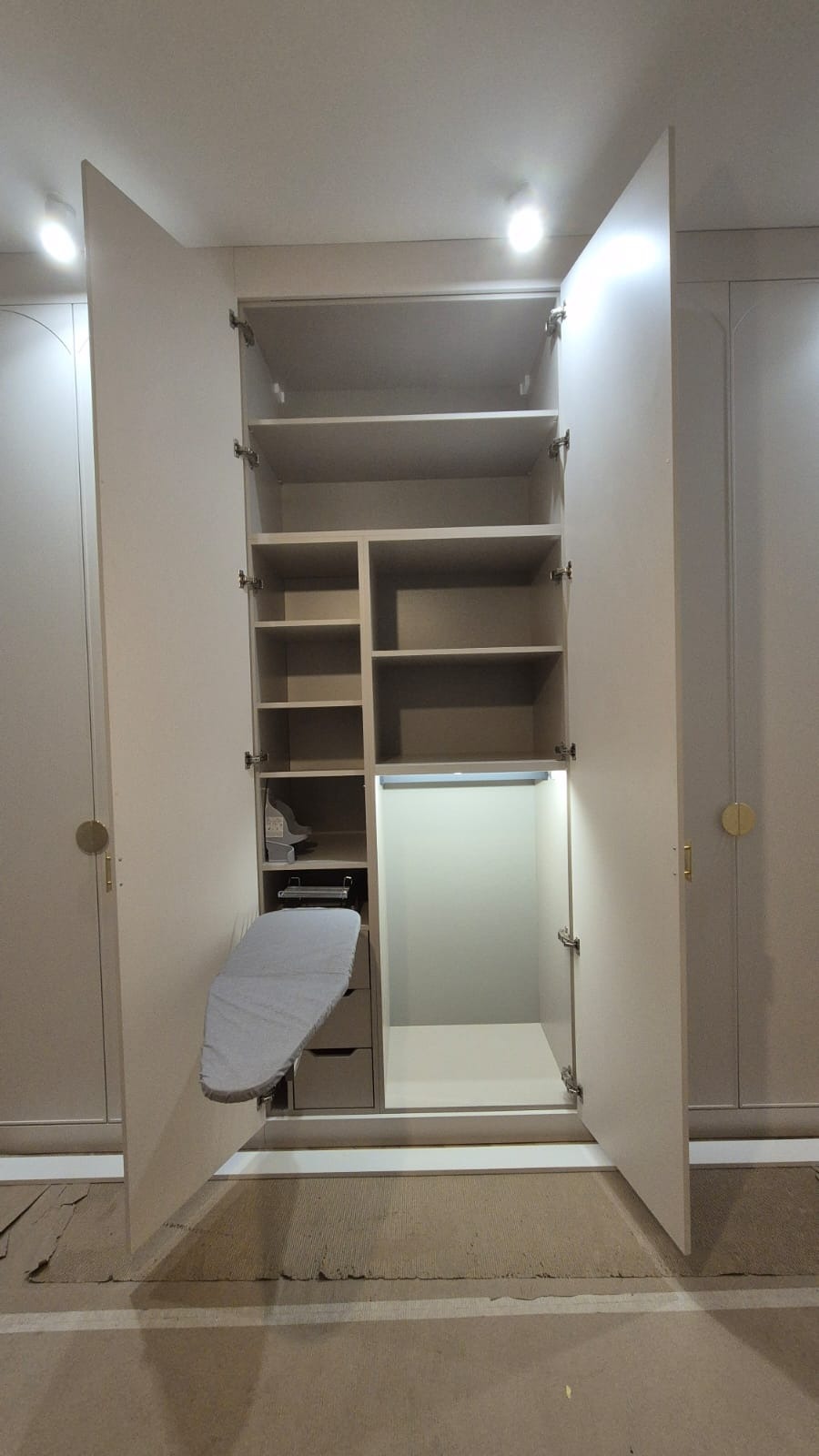 Wnętrze szafy z półkami i wysuwanym, szarym blatem do prasowania, oświetlone punktowo, na podłodze widoczne zabezpieczenia.