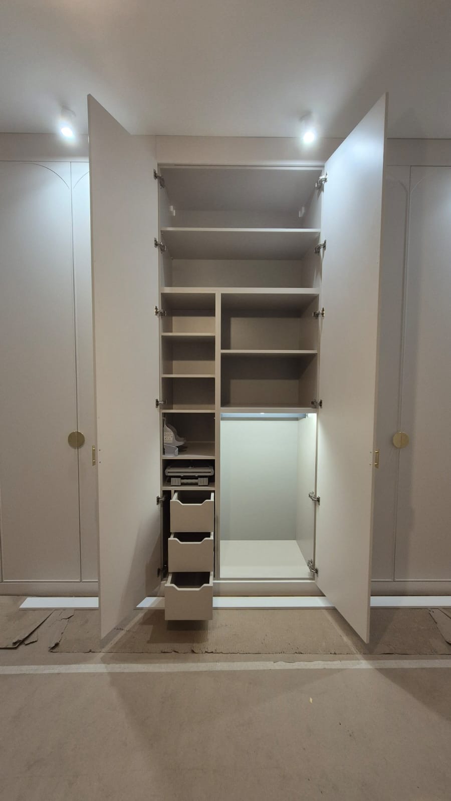 Jasna, otwarta szafa wnękowa z szufladami i półkami, oświetlenie LED wewnątrz, widoczne zawiasy i złote okrągłe uchwyty na sąsiednich drzwiach.