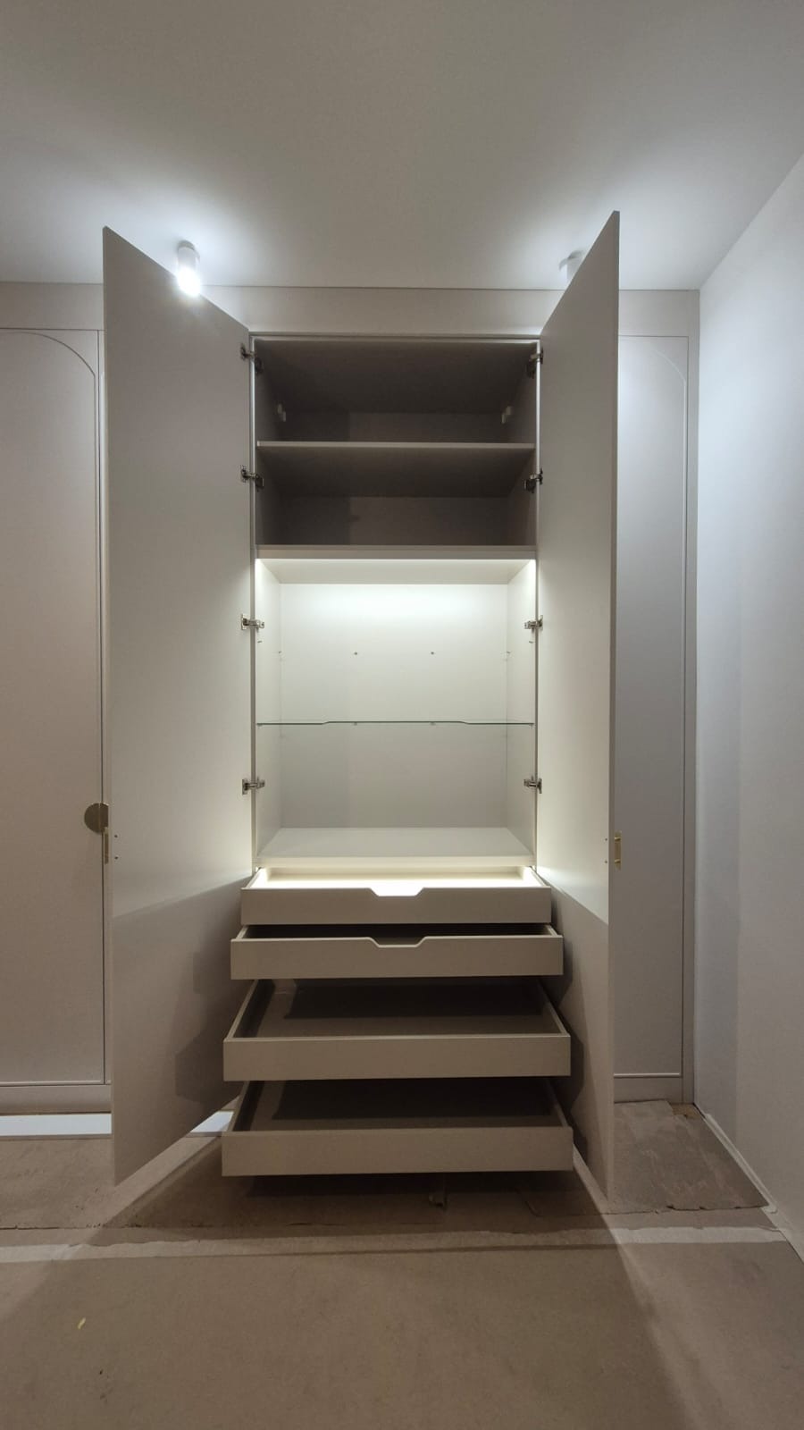 Otwarta, jasna szafa z szufladami i półkami, oświetlona od wewnątrz, z widocznymi zawiasami i złotymi uchwytami, na tle surowej podłogi w trakcie wykończenia.