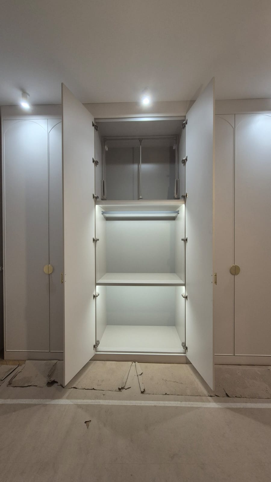 Wnętrze szafy wnękowej z otwartymi drzwiami, podświetlone półki, widoczne zawiasy i drążek, w tle zamknięte segmenty szafy z okrągłymi, złotymi uchwytami, surowa podłoga w trakcie montażu.