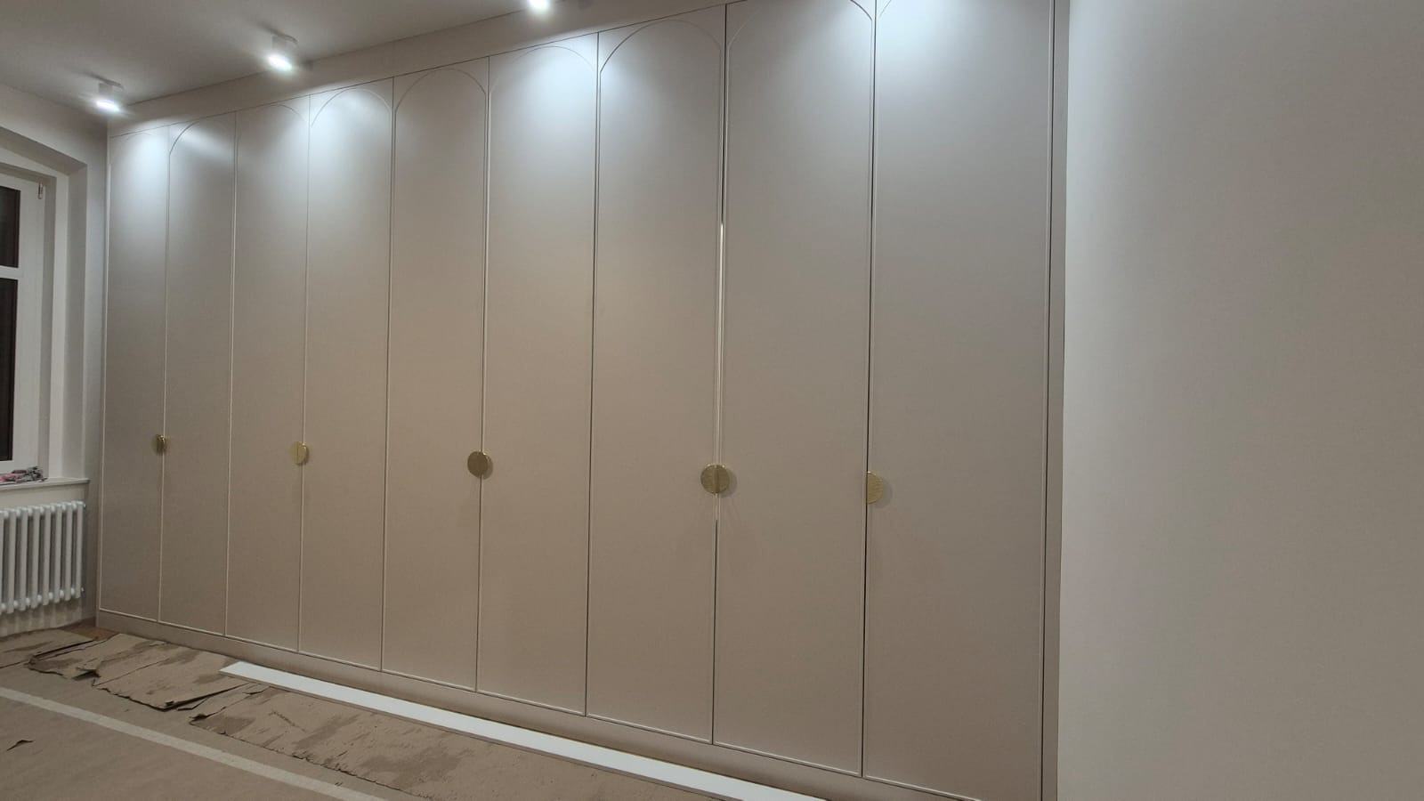 Biała, wnękowa szafa z zaokrąglonymi górnymi krawędziami drzwi i okrągłymi, złotymi uchwytami, na podłodze karton zabezpieczający.