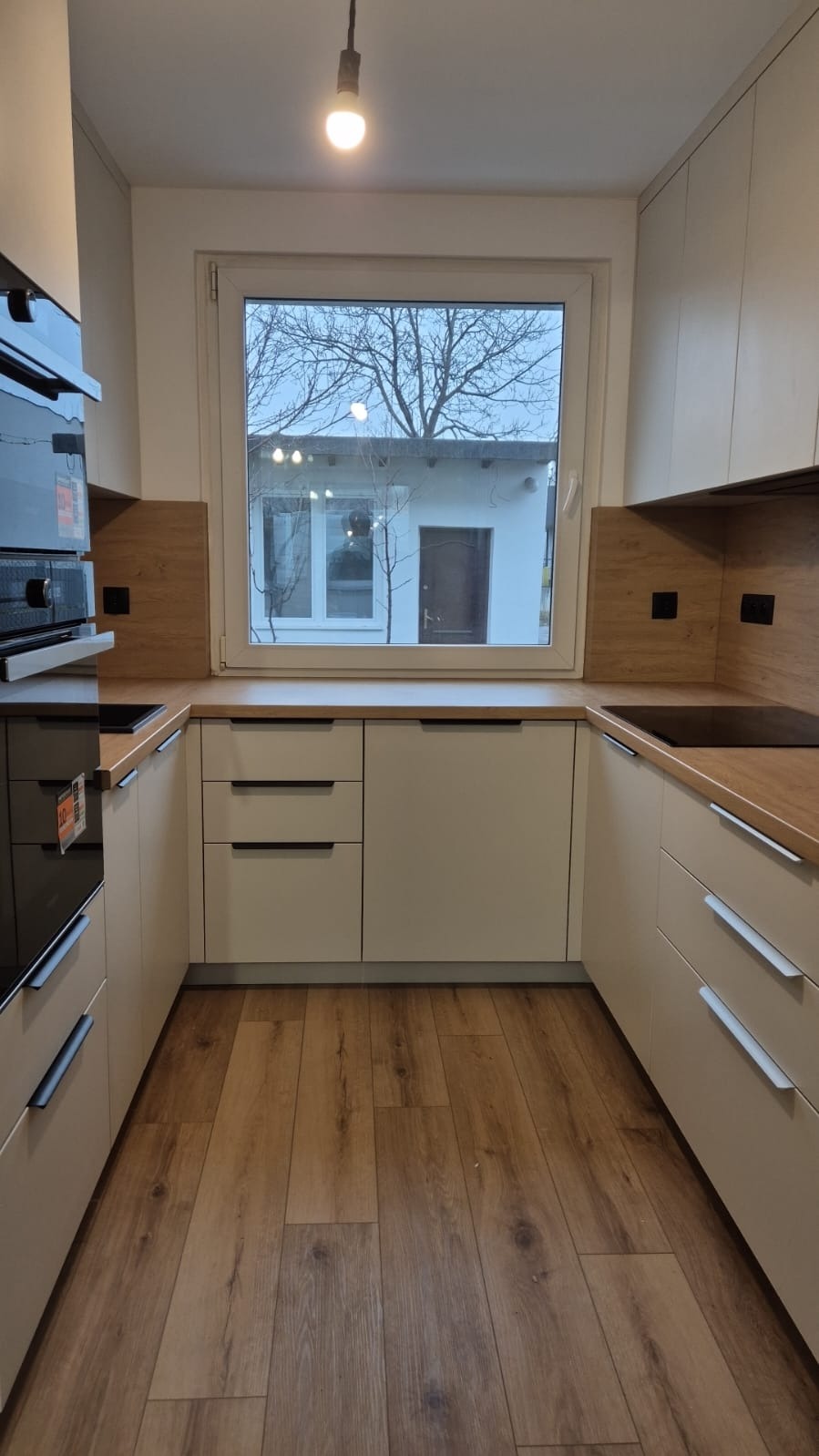 Kompaktowa kuchnia na wymiar w jasnych kolorach z drewnianym blatem i widokiem na ogród przez okno.