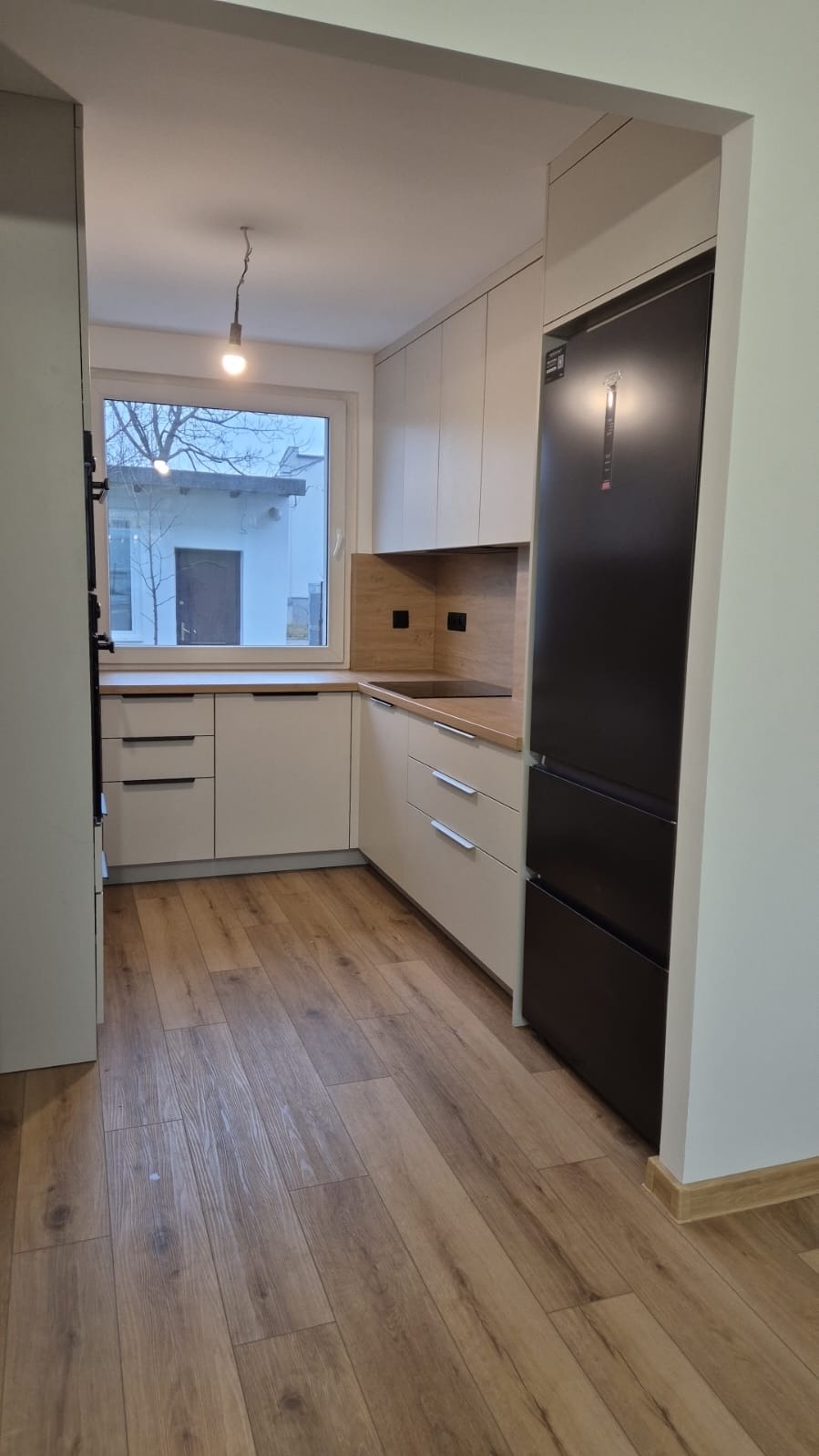 Jasna kuchnia z drewnianym blatem, czarną lodówką i drewnianą podłogą widziana z perspektywy wejścia, z oknem wychodzącym na podwórze.