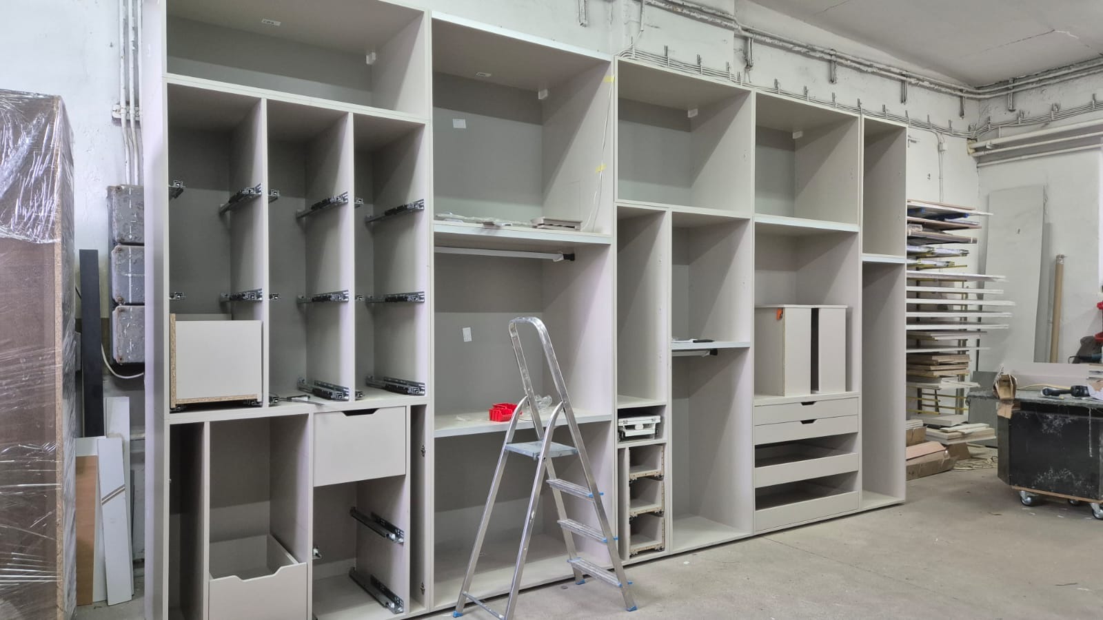 Montaż szafy wnękowej z szufladami i półkami, widoczne prowadnice, drabina aluminiowa i narzędzia w warsztacie.