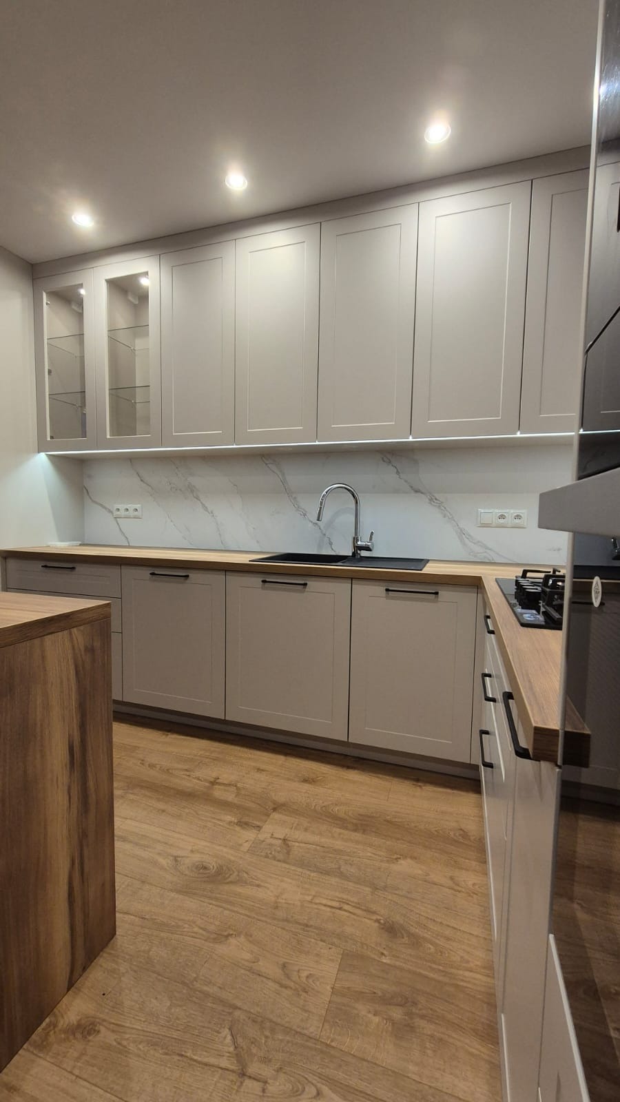 Jasnoszara zabudowa kuchenna z drewnianym blatem, białym marmurowym panelem nad blatem i oświetleniem LED podszafkowym; widoczny zlew, bateria i płyta grzewcza.