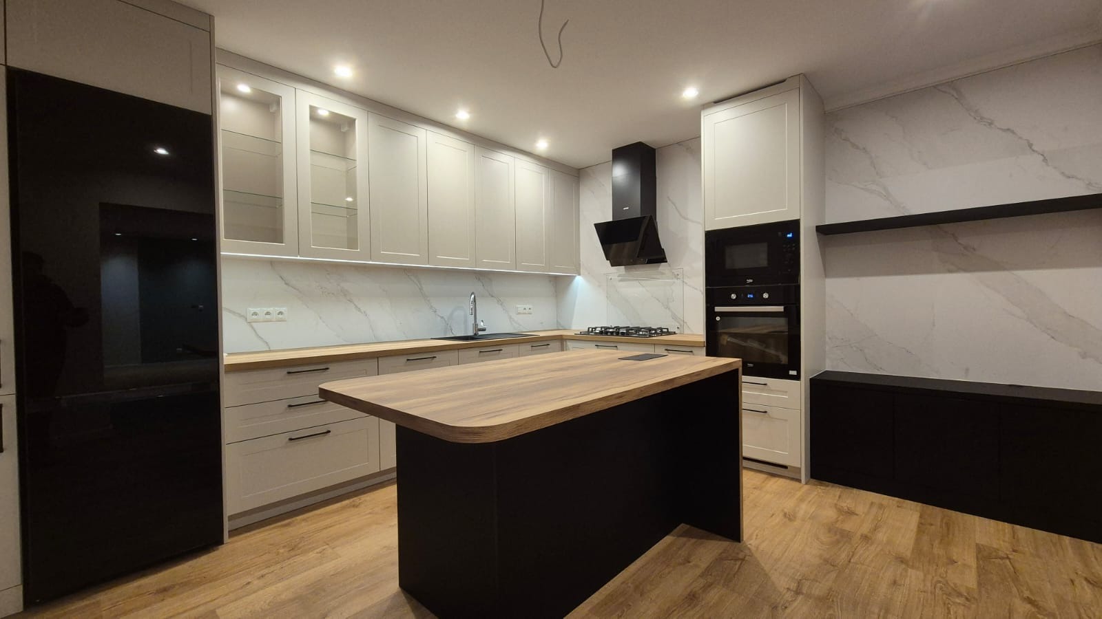 Nowoczesna kuchnia z wyspą, jasne szafki z drewnianymi blatami, czarny sprzęt AGD i marmurowe ściany.