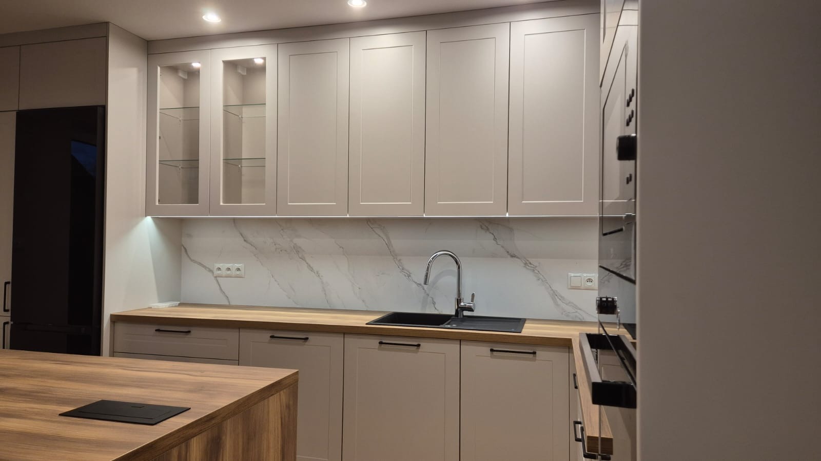 Jasna kuchnia z drewnianym blatem, marmurowym backsplashiem i szklanymi szafkami wiszącymi z oświetleniem LED pod spodem, z czarnym zlewem i baterią oraz wyspą kuchenną z gniazdem elektrycznym.