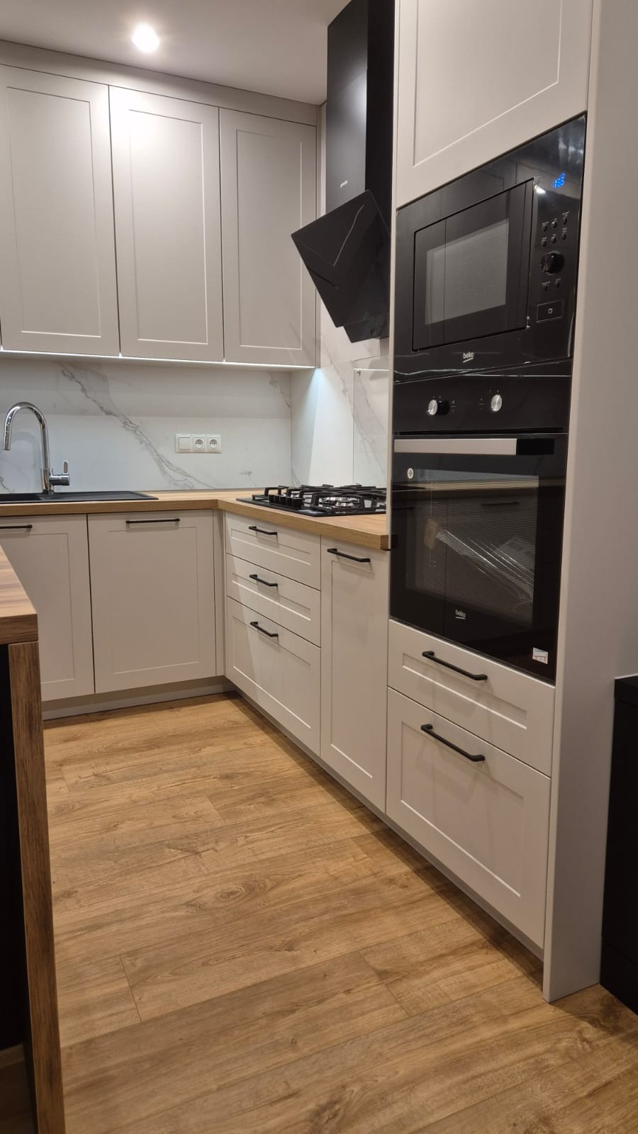 Jasnoszara zabudowa kuchenna z drewnianym blatem, czarnymi uchwytami i sprzętem AGD marki Beko: piekarnik, mikrofalówka i okap.