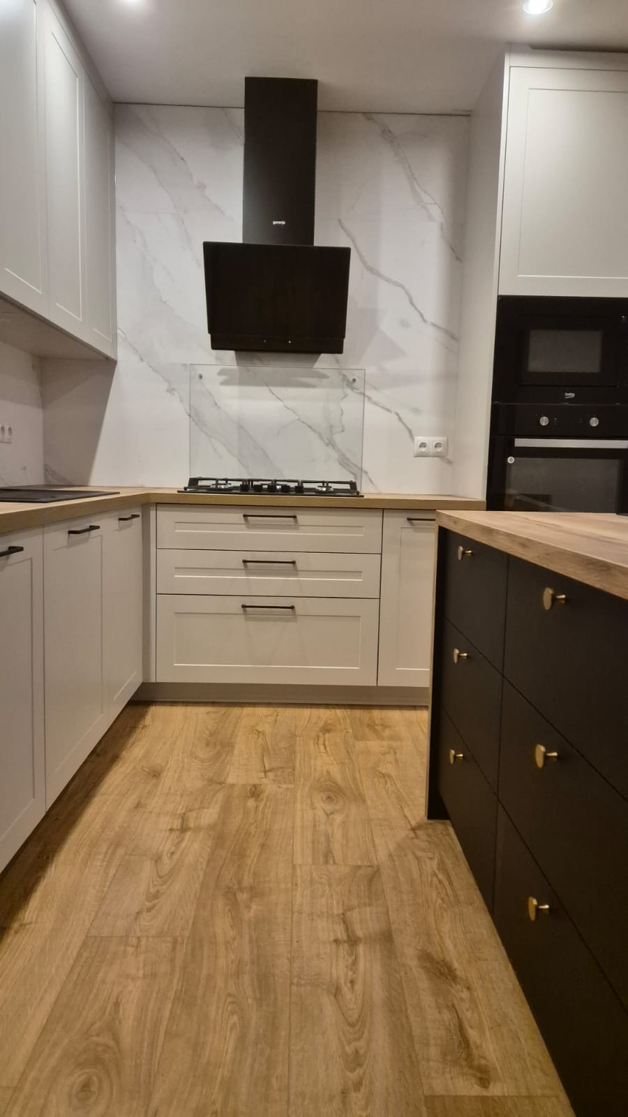 Nowoczesna kuchnia z białymi frontami szafek, drewnianym blatem i czarną wyspą, marmurowy backsplash i czarny okap.