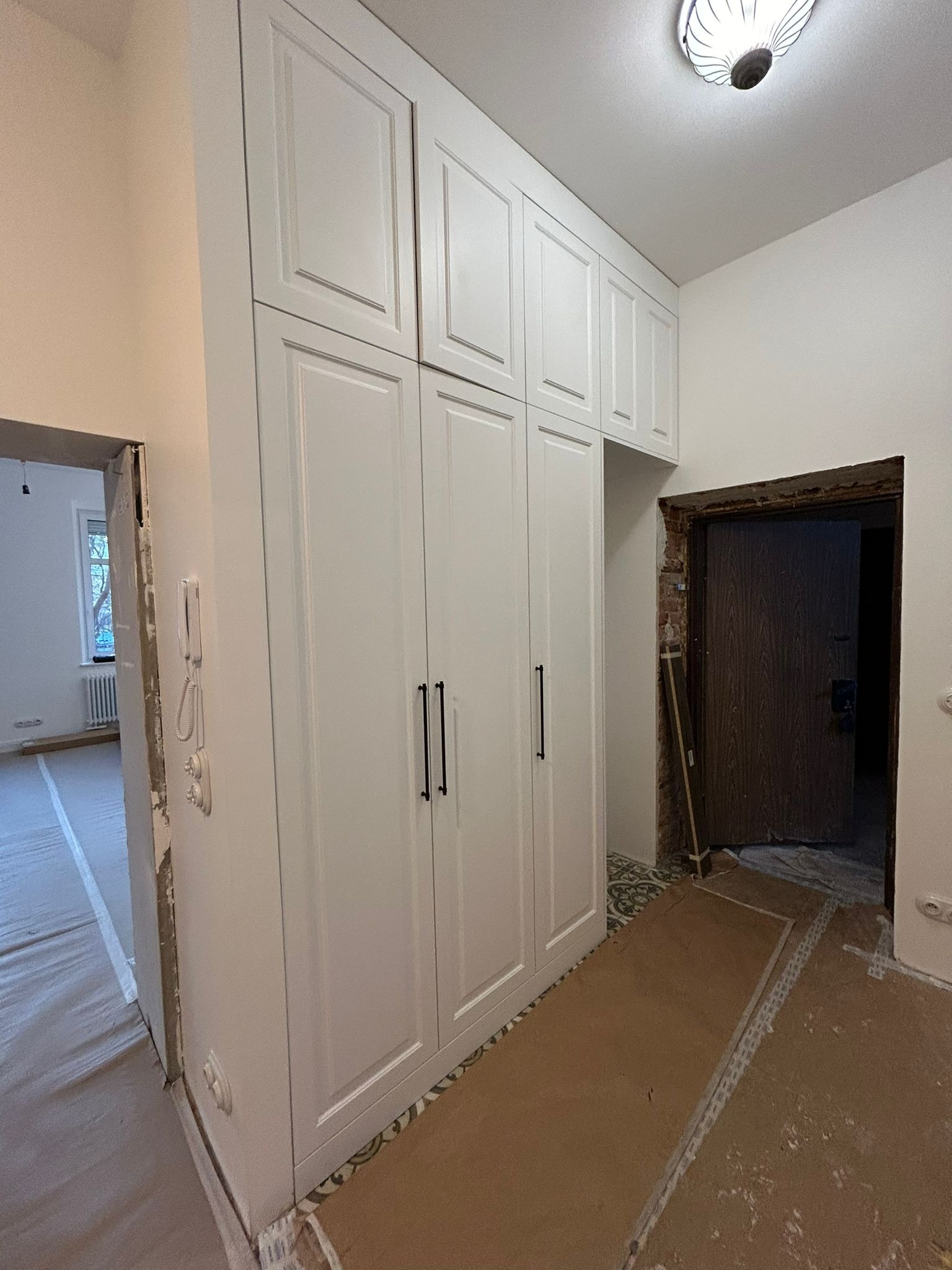 Biała, wysoka szafa z czarnymi uchwytami w trakcie montażu, widoczne zabezpieczenia podłogi i niedokończone wykończenia ścian.