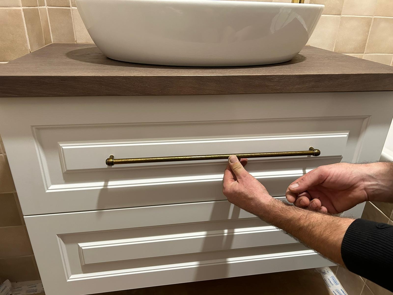 Montaż uchwytu w złotej barwie do białej szafki łazienkowej pod umywalką nablatową z blatem w kolorze drewna.