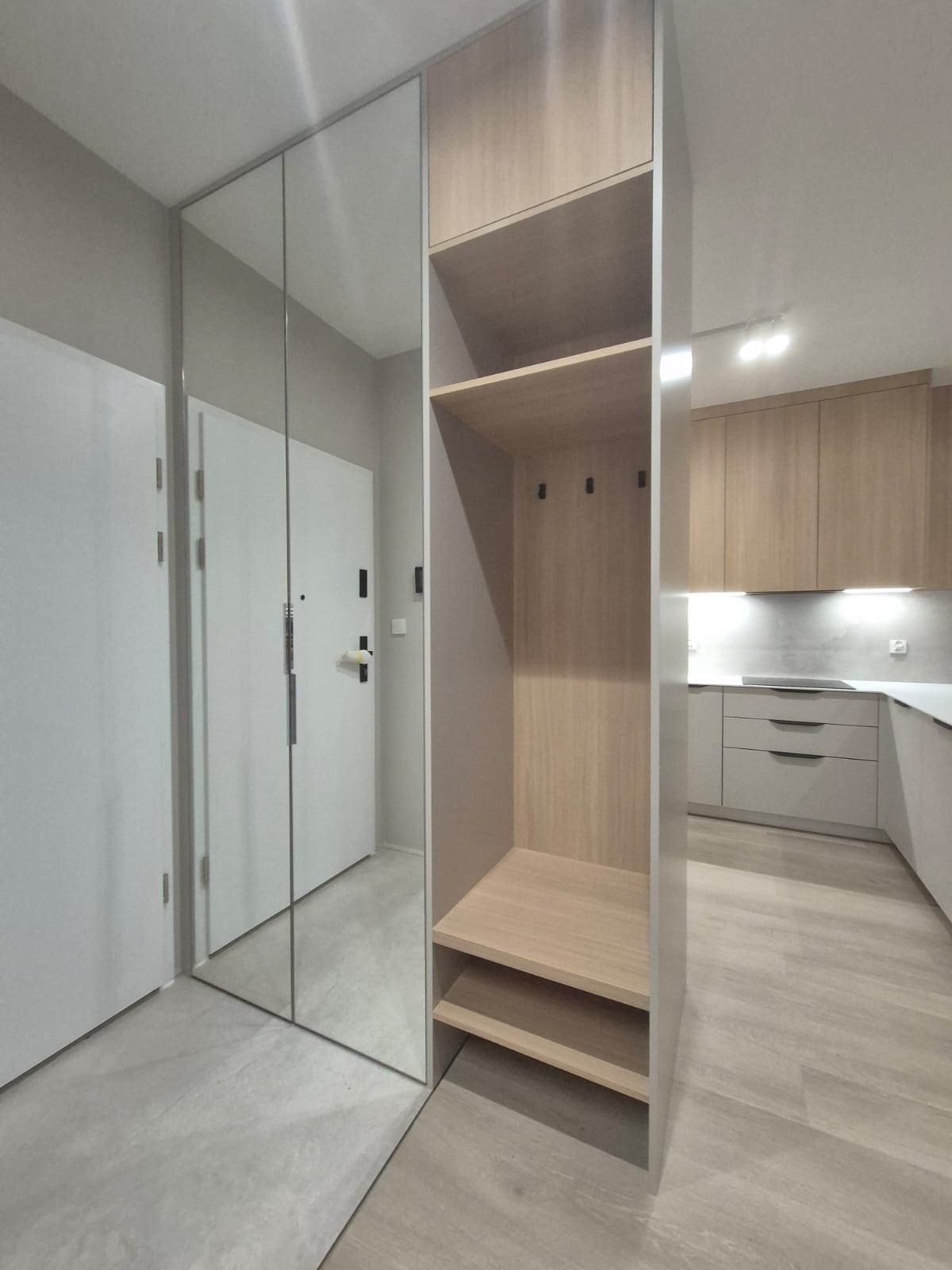 Nowoczesna szafa z lustrzanymi drzwiami i otwartą półką na buty w przedpokoju, z widocznym fragmentem kuchni w tle.