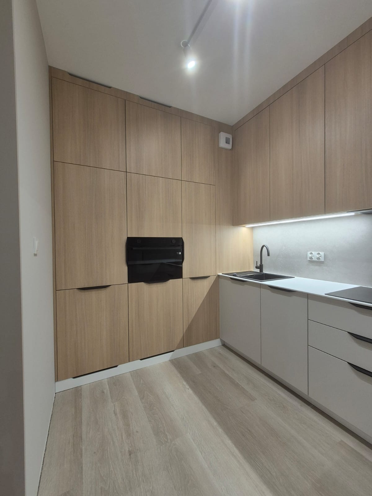 Nowoczesna kuchnia z drewnianymi frontami szafek, czarnym piekarnikiem w zabudowie i szarymi dolnymi szafkami z czarną baterią. Oświetlenie LED podszafkowe i jasna podłoga.