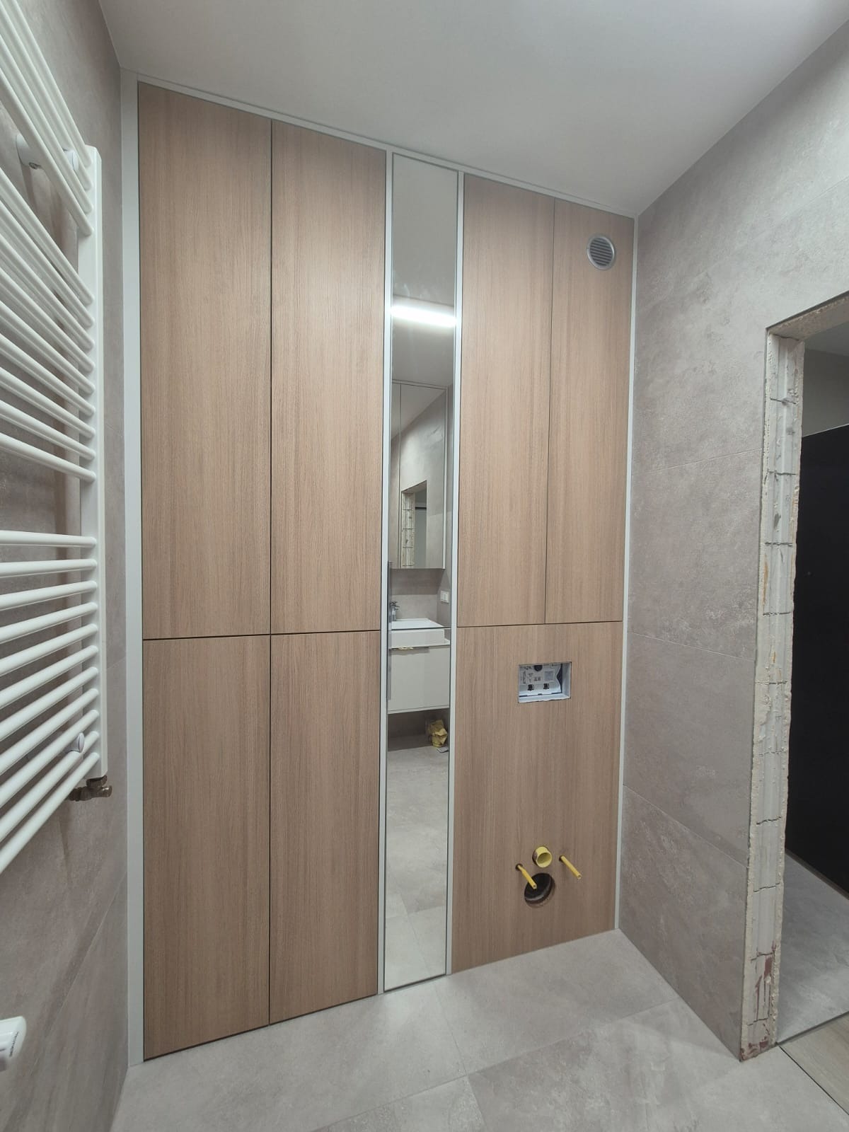 Wnętrze łazienki z zabudową meblową w odcieniach drewna z lustrem, widoczne przyłącza hydrauliczne i grzejnik drabinkowy, płytki imitujące beton na ścianach i podłodze.