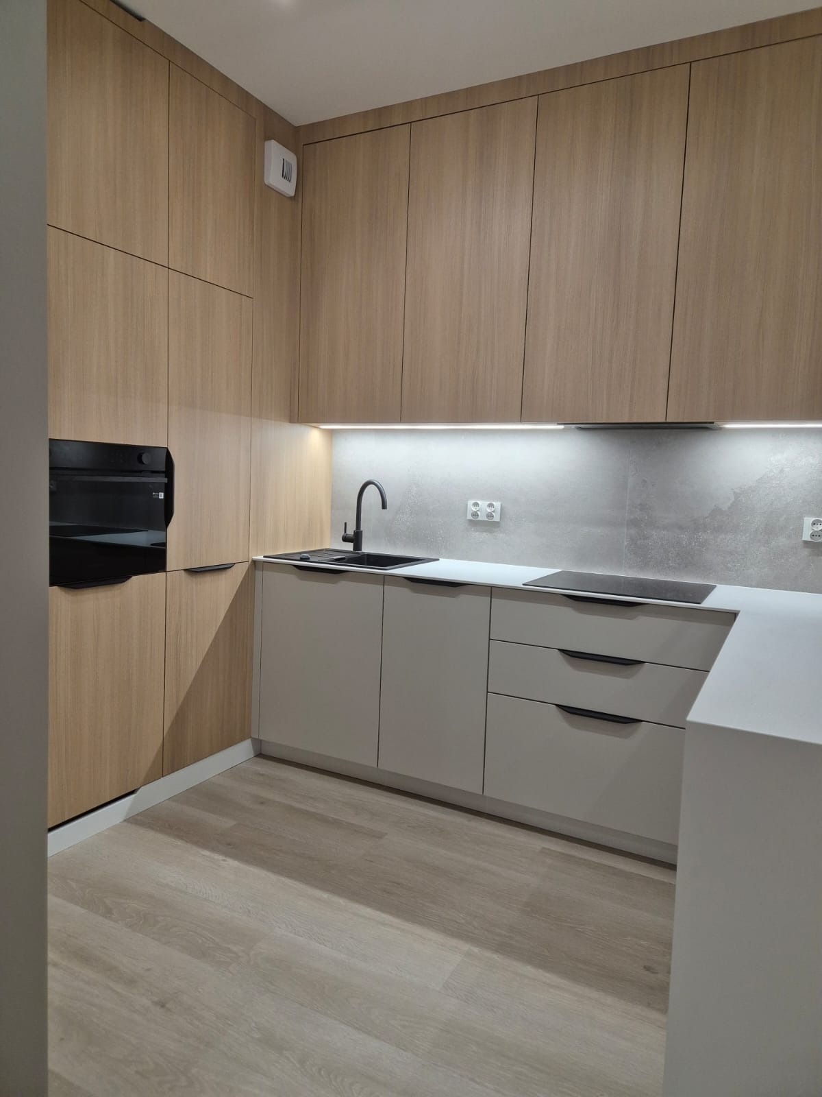 Nowoczesna kuchnia na wymiar z drewnianymi górnymi szafkami, szarymi dolnymi szafkami i czarną armaturą, oświetlona taśmą LED pod szafkami wiszącymi.