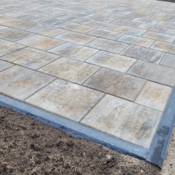 JABUD Roboty ziemne - Nowo ułożona, beżowo-szara kostka brukowa z ciemnoszarą obwódką, granicząca z przygotowanym podłożem z ziemi ogrodowej. W tle widoczny metalowy płot i dom.