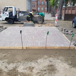 JABUD Roboty ziemne - Prace brukarskie w toku: przygotowane podłoże z piasku, częściowo ułożona kostka w odcieniach czerwieni i szarości, ekipa budowlana przy ciężarówce, w tle budynek z czerwonej cegły i niebieski...