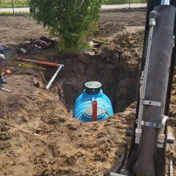 JABUD Roboty ziemne - Niebieski zbiornik zakopany w ziemi w wykopie, widoczny fragment koparki i rury, drzewo w tle.