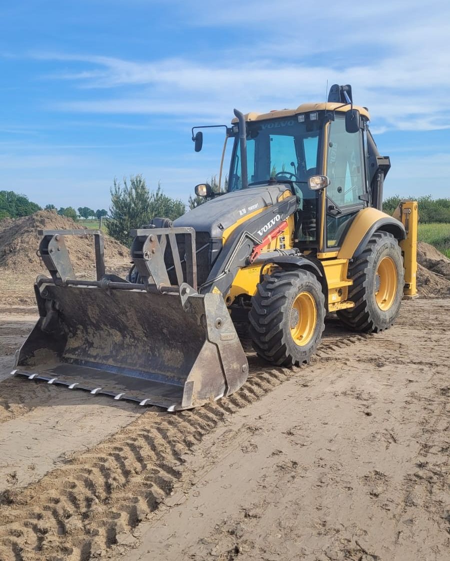 Żółto-szara koparko-ładowarka Volvo z łyżką, stojąca na piaszczystym terenie z widocznymi śladami opon, na tle błękitnego nieba z delikatnymi chmurami i zielenią w oddali.