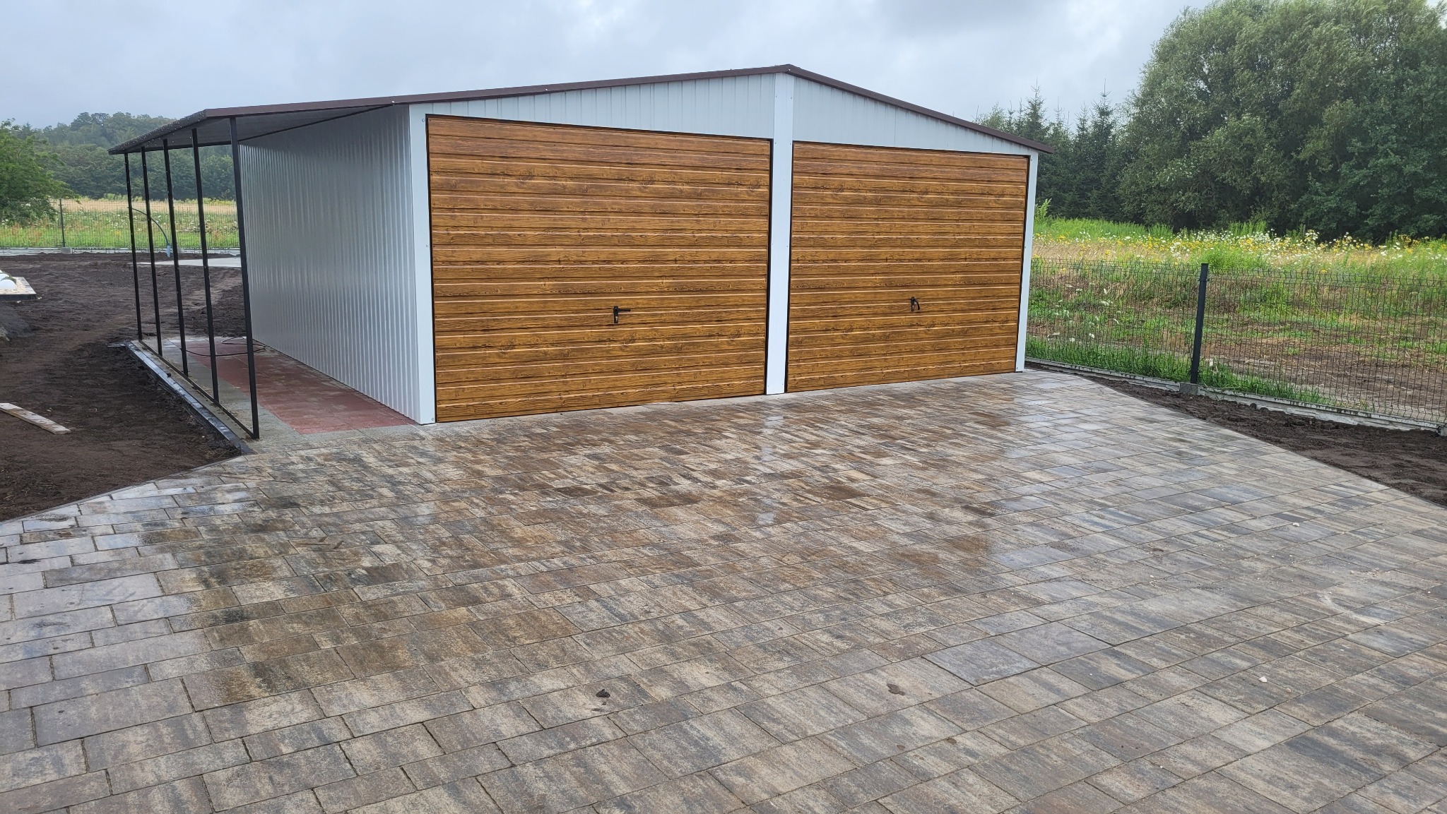 Nowo ułożona, wilgotna kostka brukowa w odcieniach szarości i brązu prowadząca do dwustanowiskowego, blaszanego garażu z bramami imitującymi drewno, widok na teren posesji z ogrodzeniem w tle.