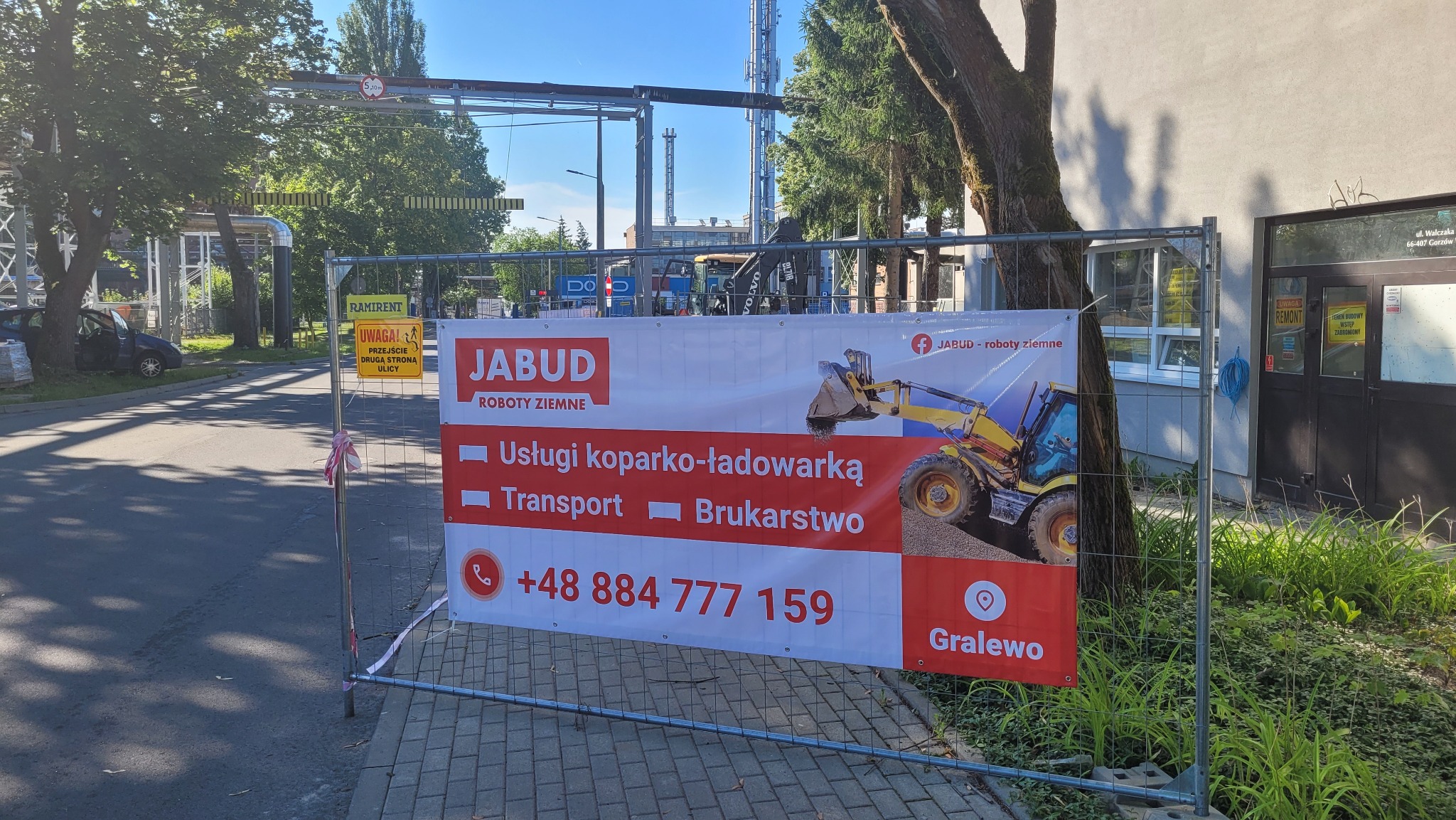 Baner reklamowy firmy JABUD Roboty Ziemne oferującej usługi koparko-ładowarką, transport i brukarstwo, z ilustracją koparki, umieszczony na ogrodzeniu budowlanym przy ulicy, w tle budynek i drzewa.