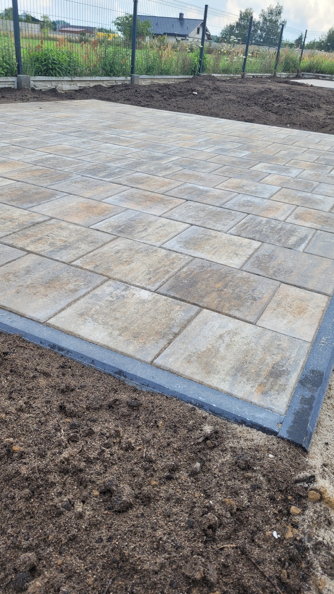 Nowo ułożona, beżowo-szara kostka brukowa z ciemnoszarą obwódką, granicząca z przygotowanym podłożem z ziemi ogrodowej. W tle widoczny metalowy płot i dom.