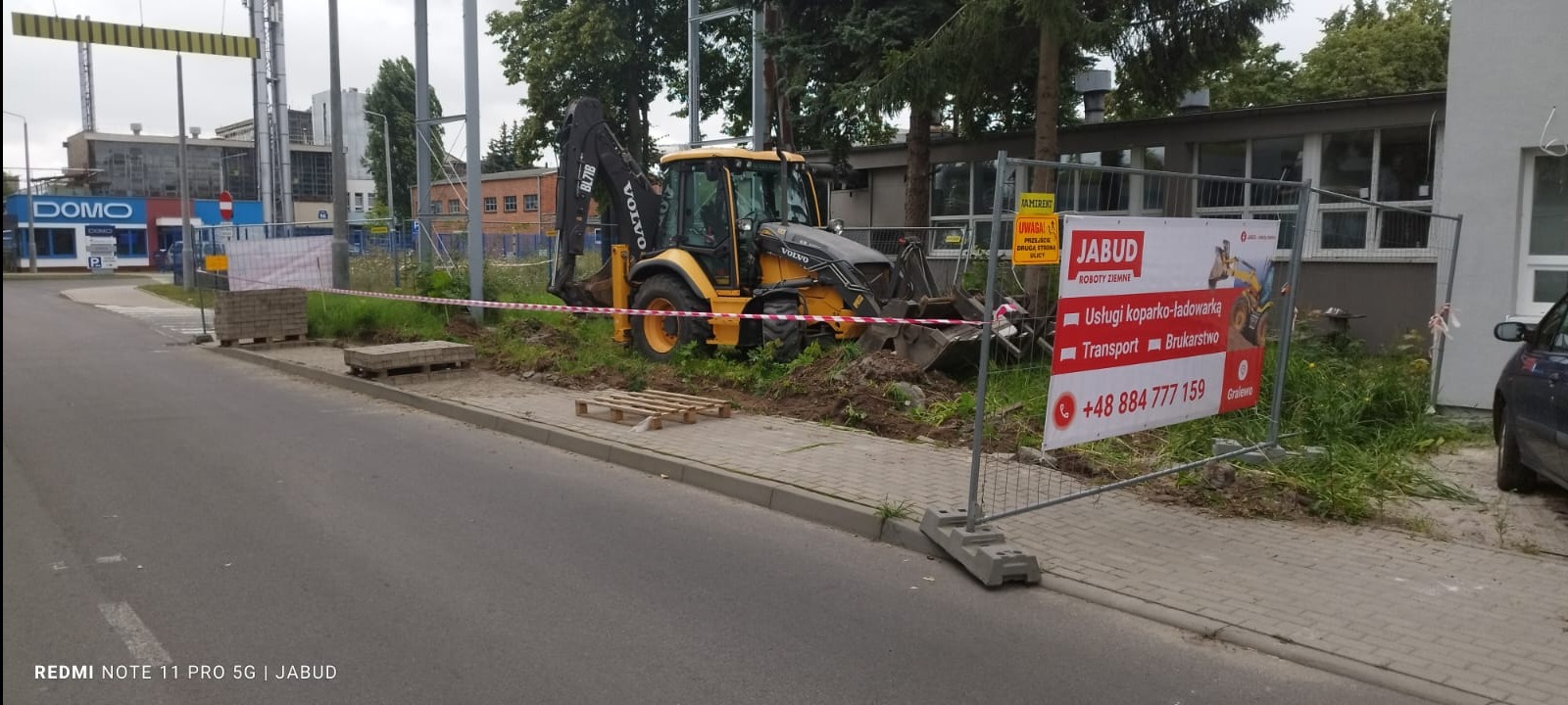 Żółta koparko-ładowarka Volvo BL71B w trakcie prac ziemnych przy chodniku, widoczny wykop, paleta i kostka brukowa, ogrodzenie z tablicą reklamową firmy JABUD oferującej usługi koparko-ładowarką...