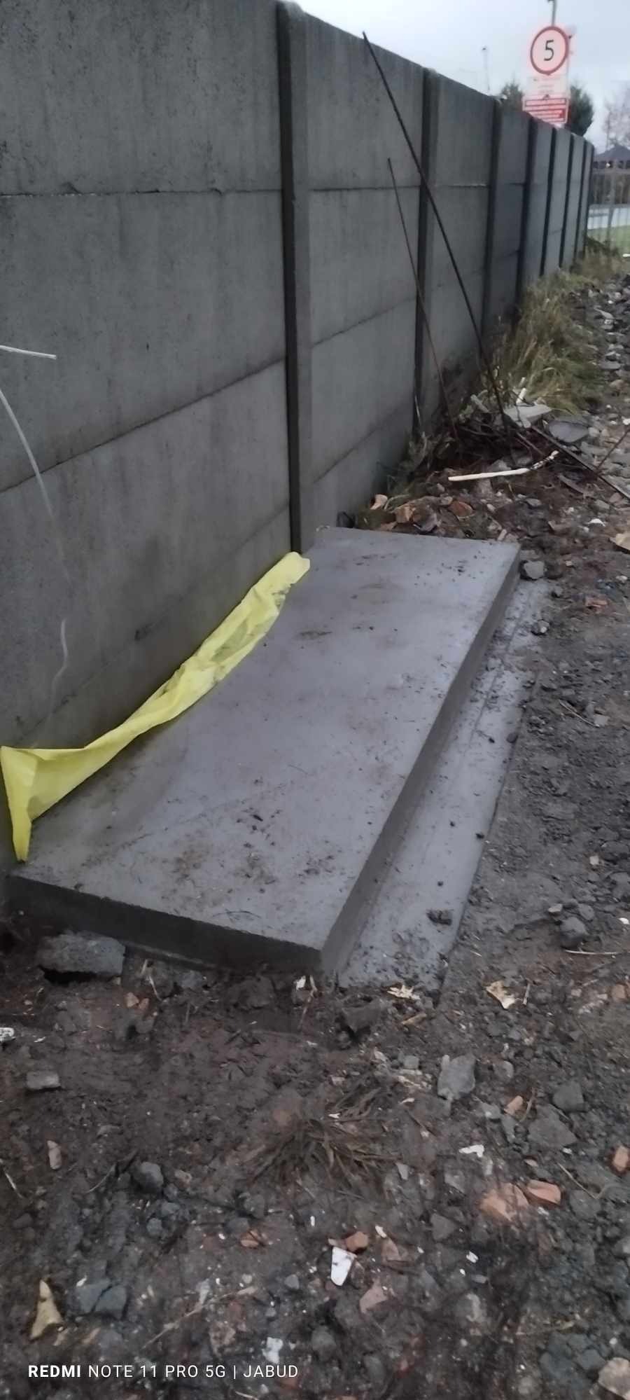 Betonowy fundament pod ogrodzenie panelowe, częściowo zabezpieczony żółtą folią, obok grząski teren.