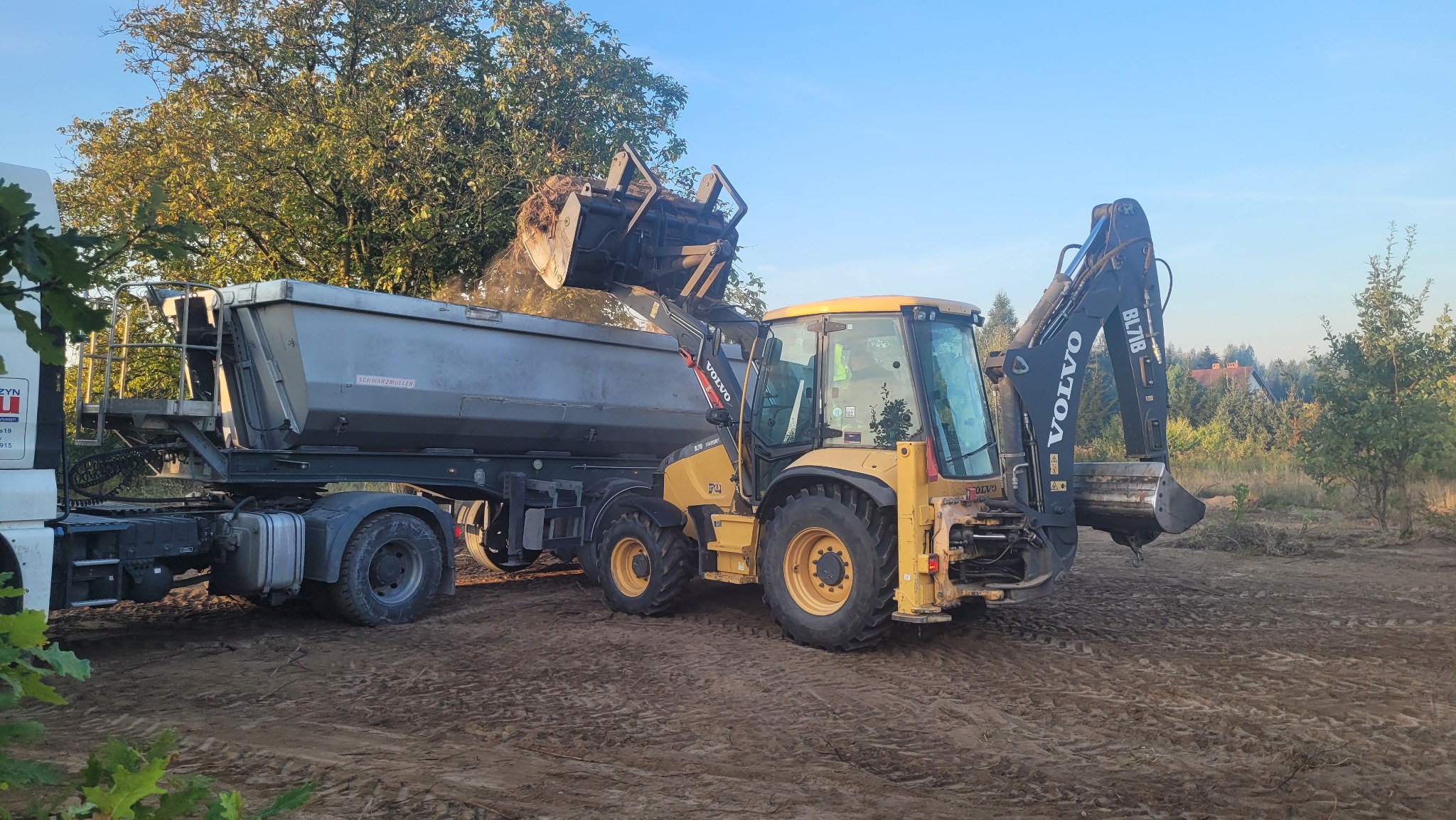 Żółta koparko-ładowarka Volvo BL71B z pełną łyżką ziemi wysypuje urobek do przyczepy ciężarówki na przygotowanym, wyrównanym terenie.