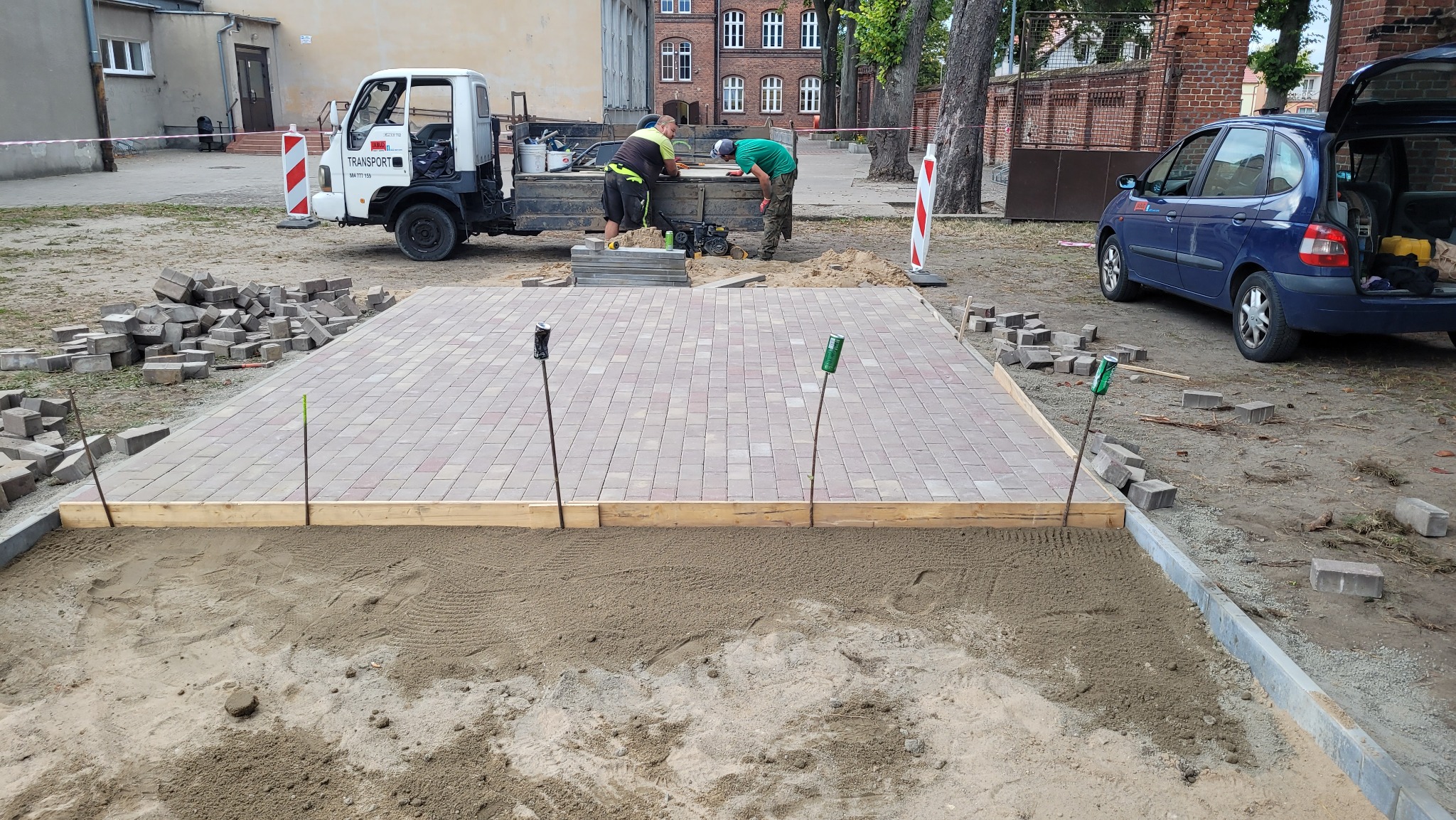 Prace brukarskie w toku: przygotowane podłoże z piasku, częściowo ułożona kostka w odcieniach czerwieni i szarości, ekipa budowlana przy ciężarówce, w tle budynek z czerwonej cegły i niebieski...