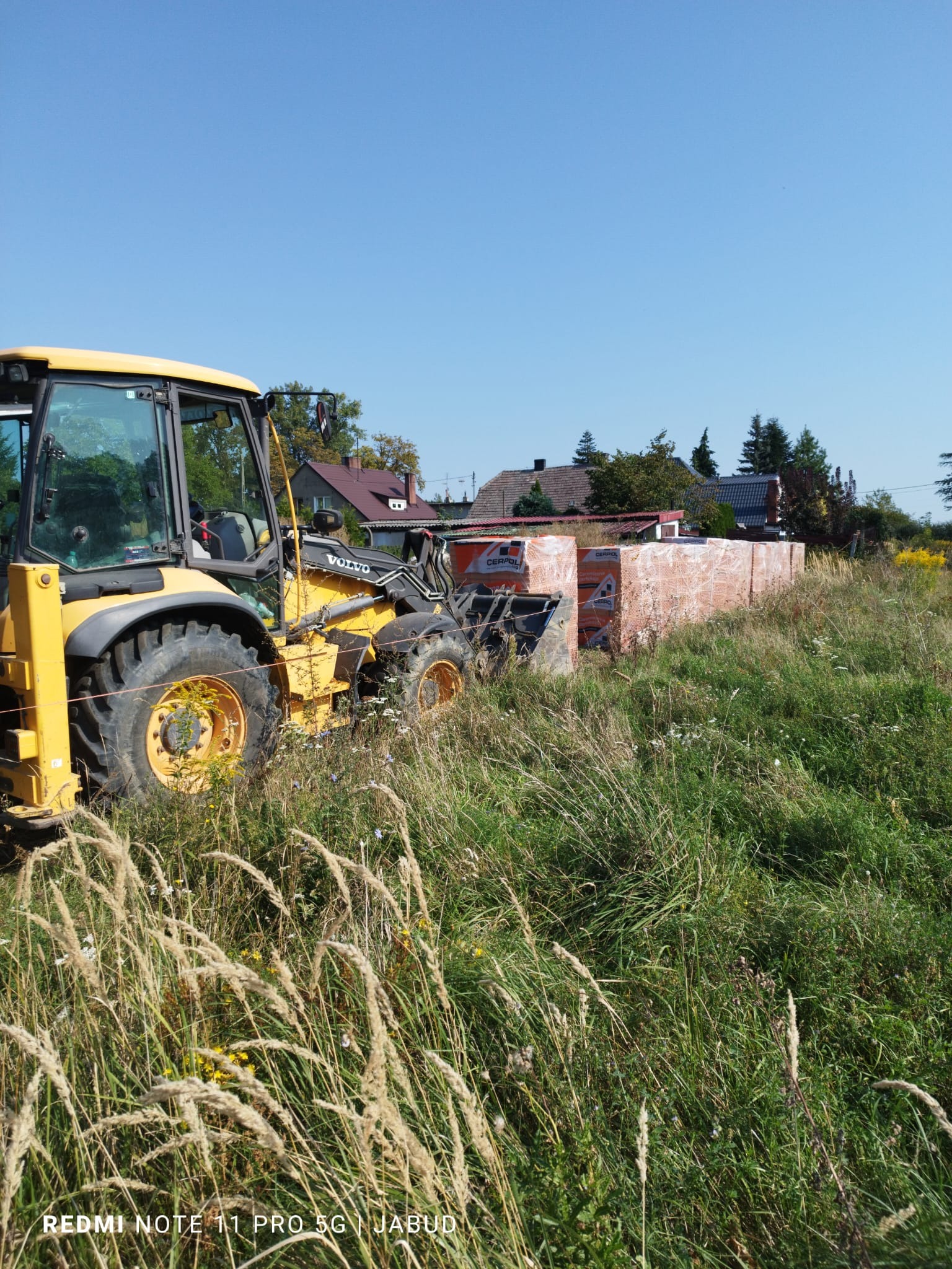 Żółta ładowarka Volvo na zarośniętym terenie obok palet z cegłami Cerpol, przygotowująca grunt pod budowę.