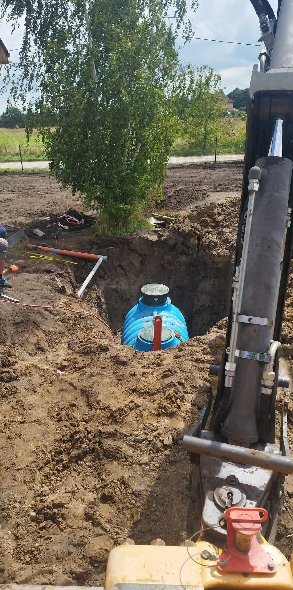 Niebieski zbiornik zakopany w ziemi w wykopie, widoczny fragment koparki i rury, drzewo w tle.