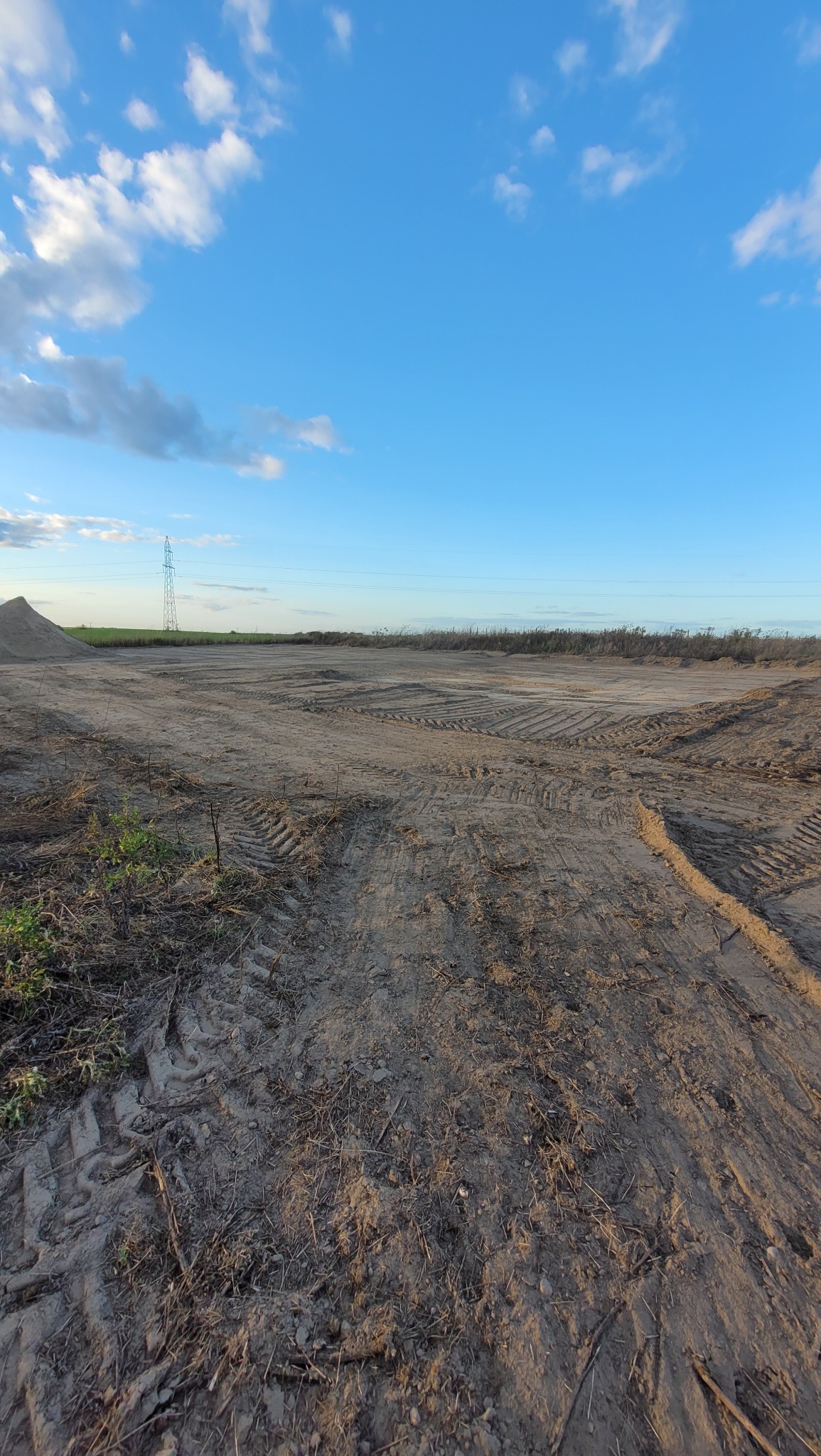Przygotowany teren pod budowę z widocznymi śladami opon, w tle słup wysokiego napięcia i błękitne niebo z chmurami.