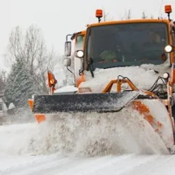 Pomarańczowy pług śnieżny w akcji, usuwający śnieg z drogi w pochmurny dzień, z widocznymi drzewami i domem w tle.