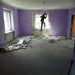Pokój w trakcie demontażu: fioletowe ściany, gruz na podłodze, pracownik na drabinie przy oknie, częściowo usunięte tynki.