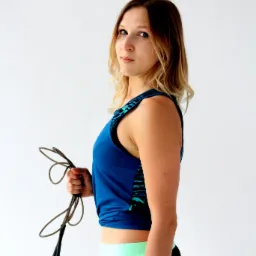 Kobieta w sportowym stroju, trzymająca skakankę, spogląda przez ramię na jasnym tle.