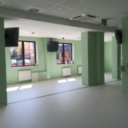 Świeżo wyremontowane biuro w odcieniach zieleni z kolumnami, nową podłogą z zaznaczoną linią, grzejnikami pod oknami i zamontowanymi, ale jeszcze ofoliowanymi telewizorami.