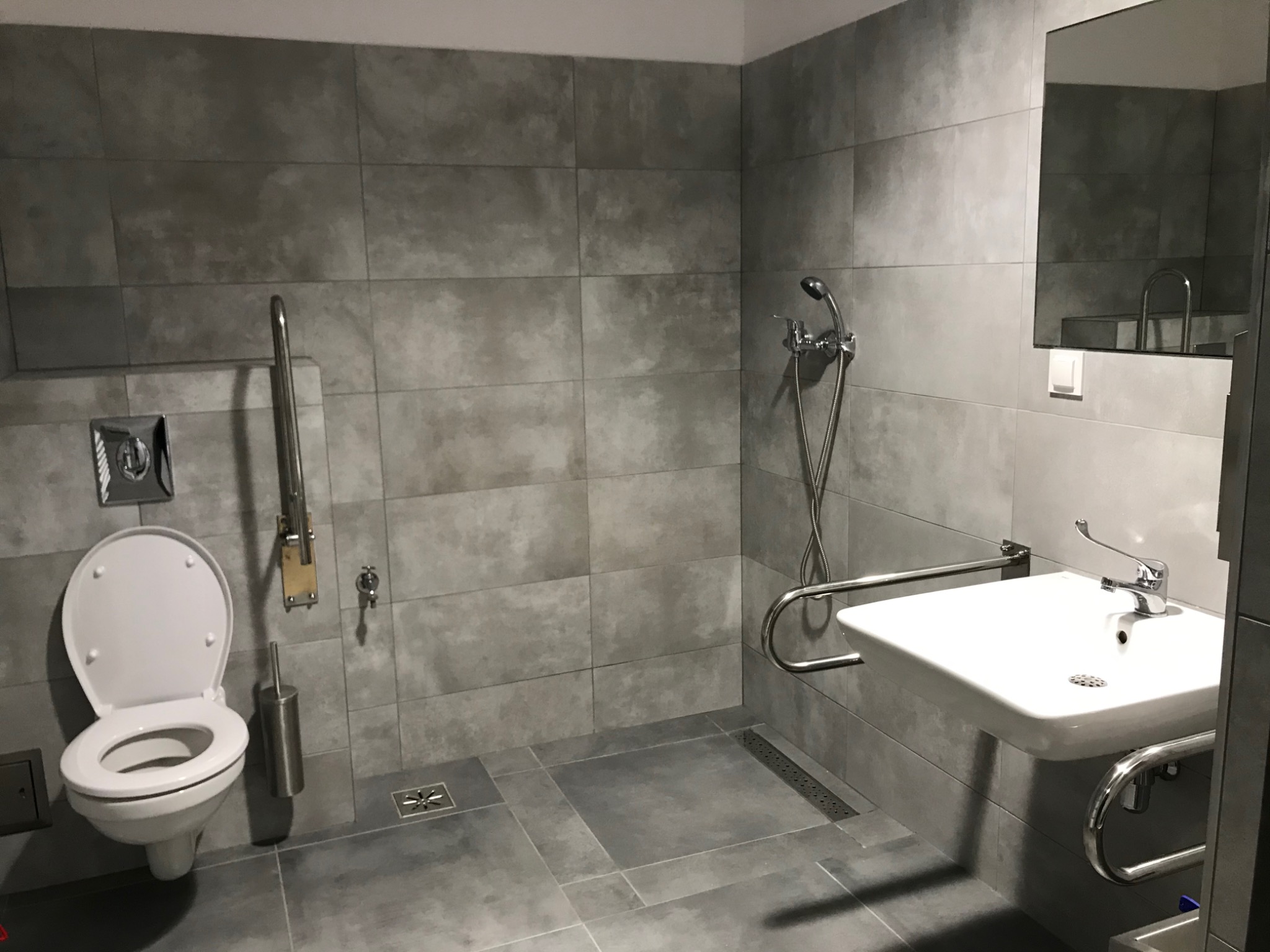 Szara łazienka z płytkami imitującymi beton, wyposażona w toaletę, umywalkę z uchwytami dla niepełnosprawnych oraz prysznic z deszczownicą i odpływem liniowym.