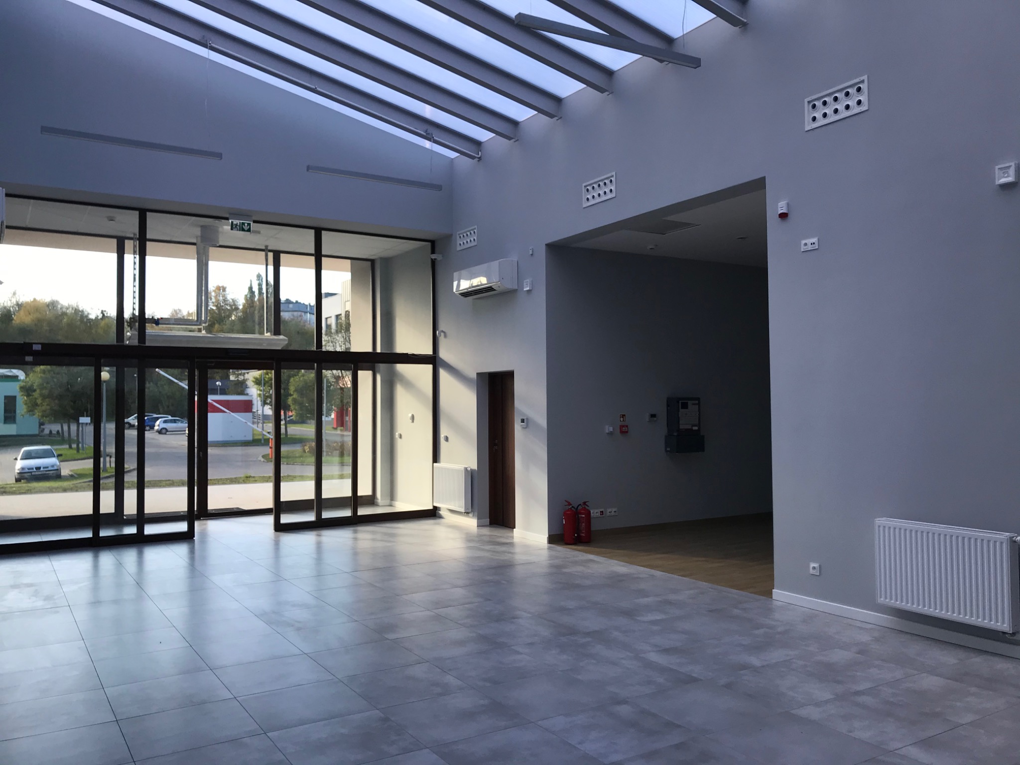 Przestronne, nowocześnie wykończone wnętrze biurowe z szarymi płytkami podłogowymi, przeszklonym wejściem i widocznym systemem wentylacji na ścianie.