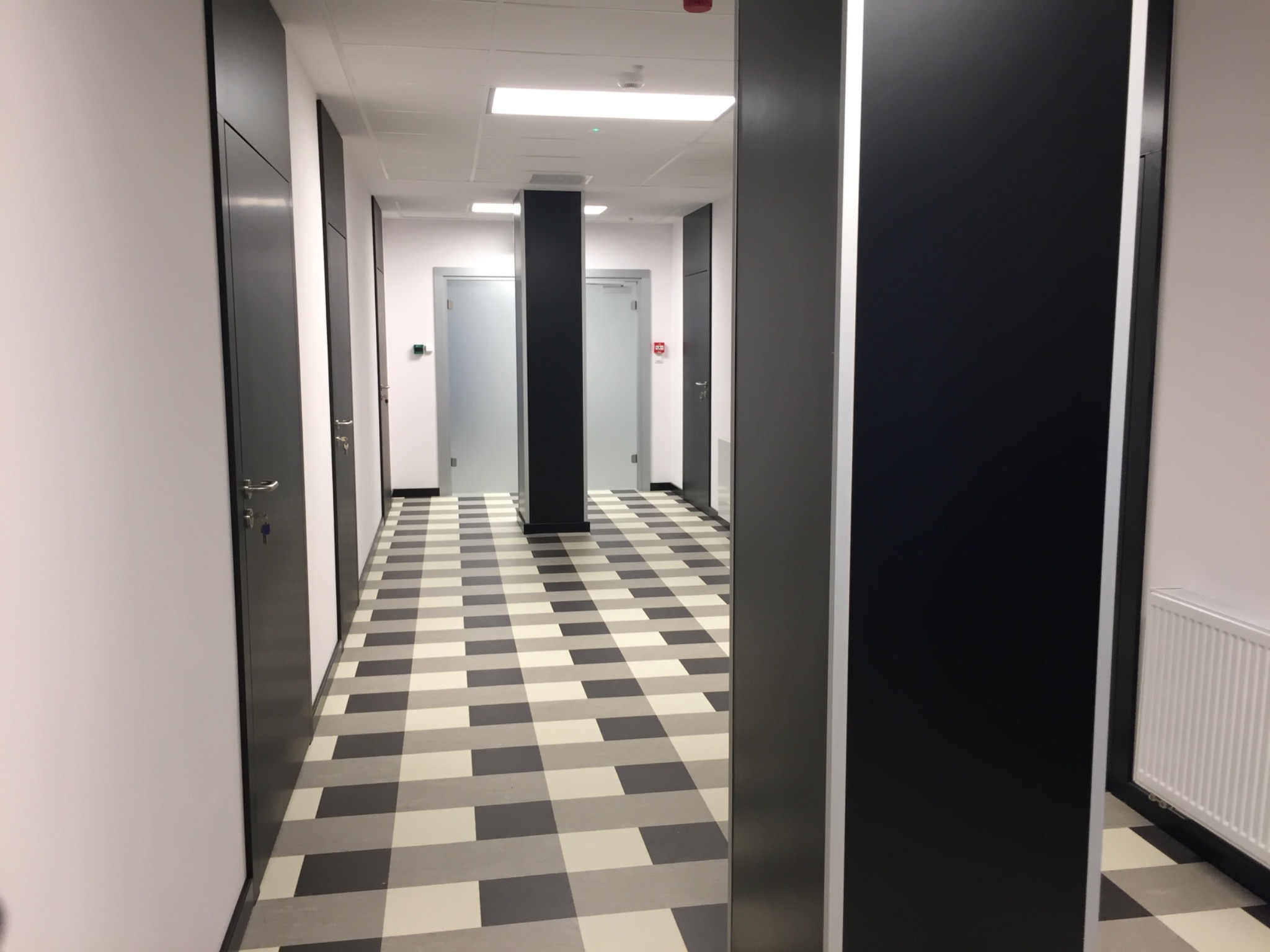 Korytarz biurowy z szachownicową podłogą, ciemnymi drzwiami i kolumnami, oświetleniem sufitowym i grzejnikiem ściennym.