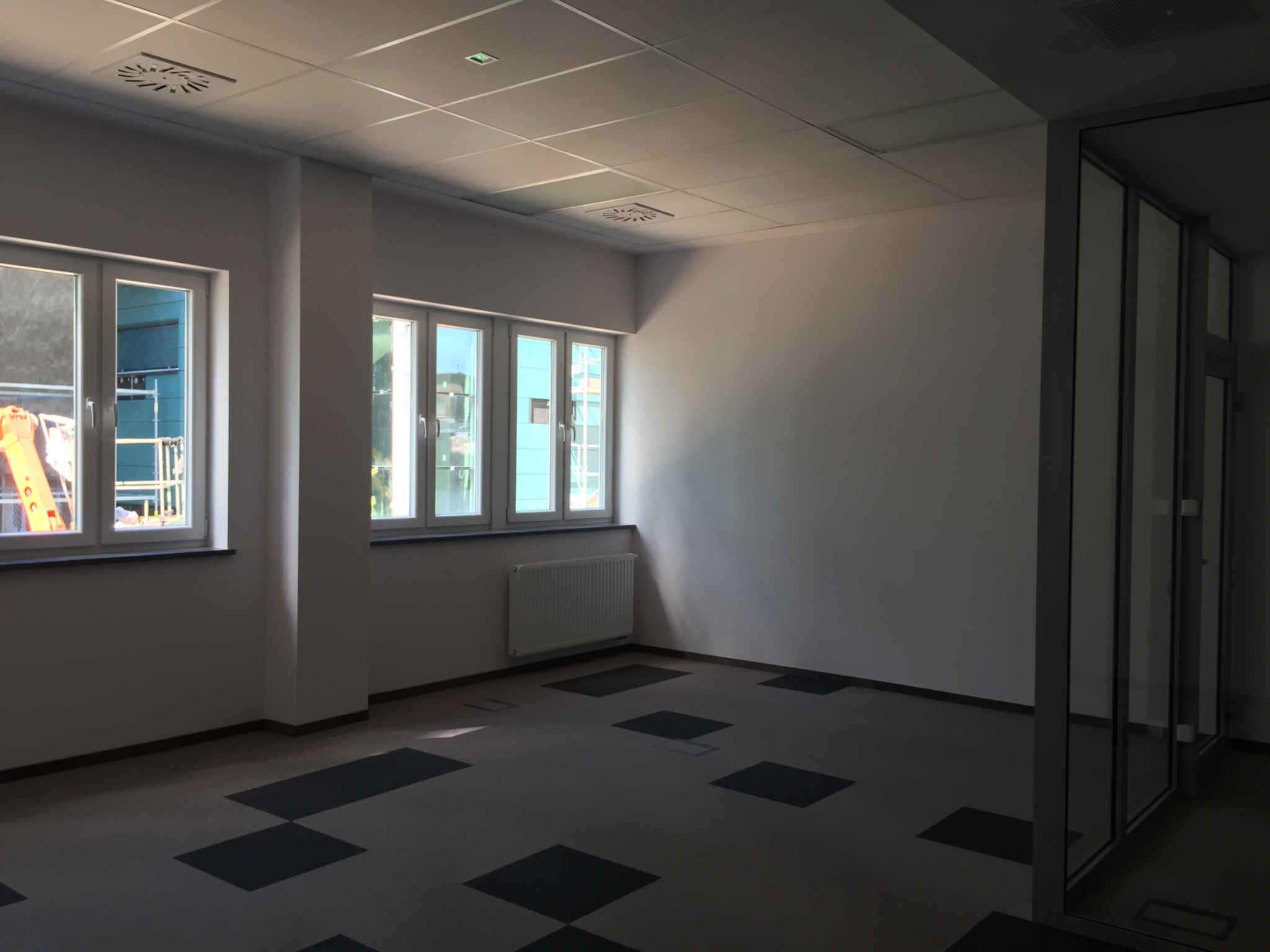 Wnętrze biura po remoncie: widok na pokój z nowymi oknami, grzejnikiem, podwieszanym sufitem z kratką wentylacyjną i podłogą w ciemne kwadraty, częściowo oszklona ściana działowa.