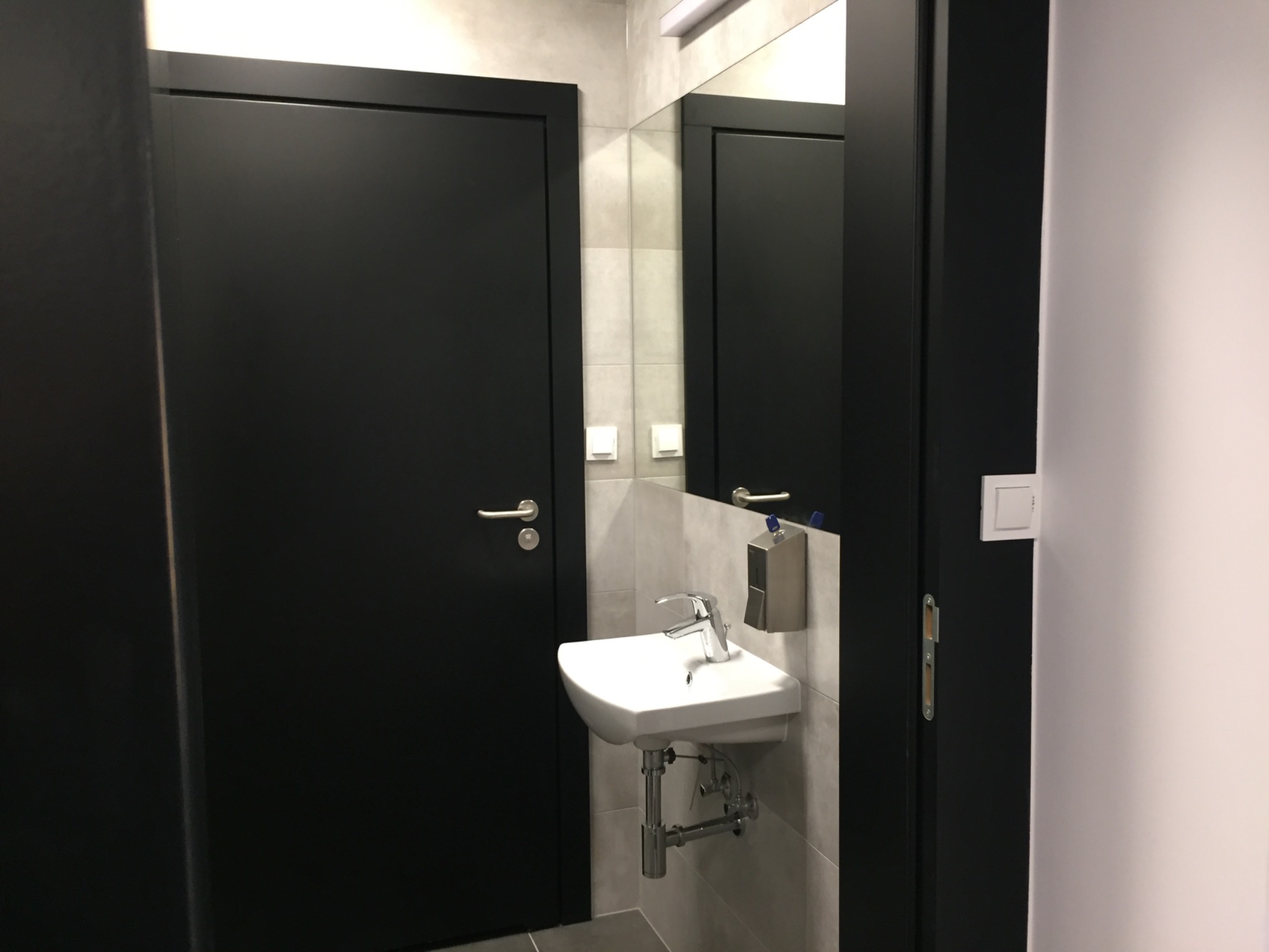 Nowoczesna łazienka biurowa z czarnymi drzwiami i białą umywalką wiszącą na szarych płytkach, widoczne metalowe elementy instalacji.