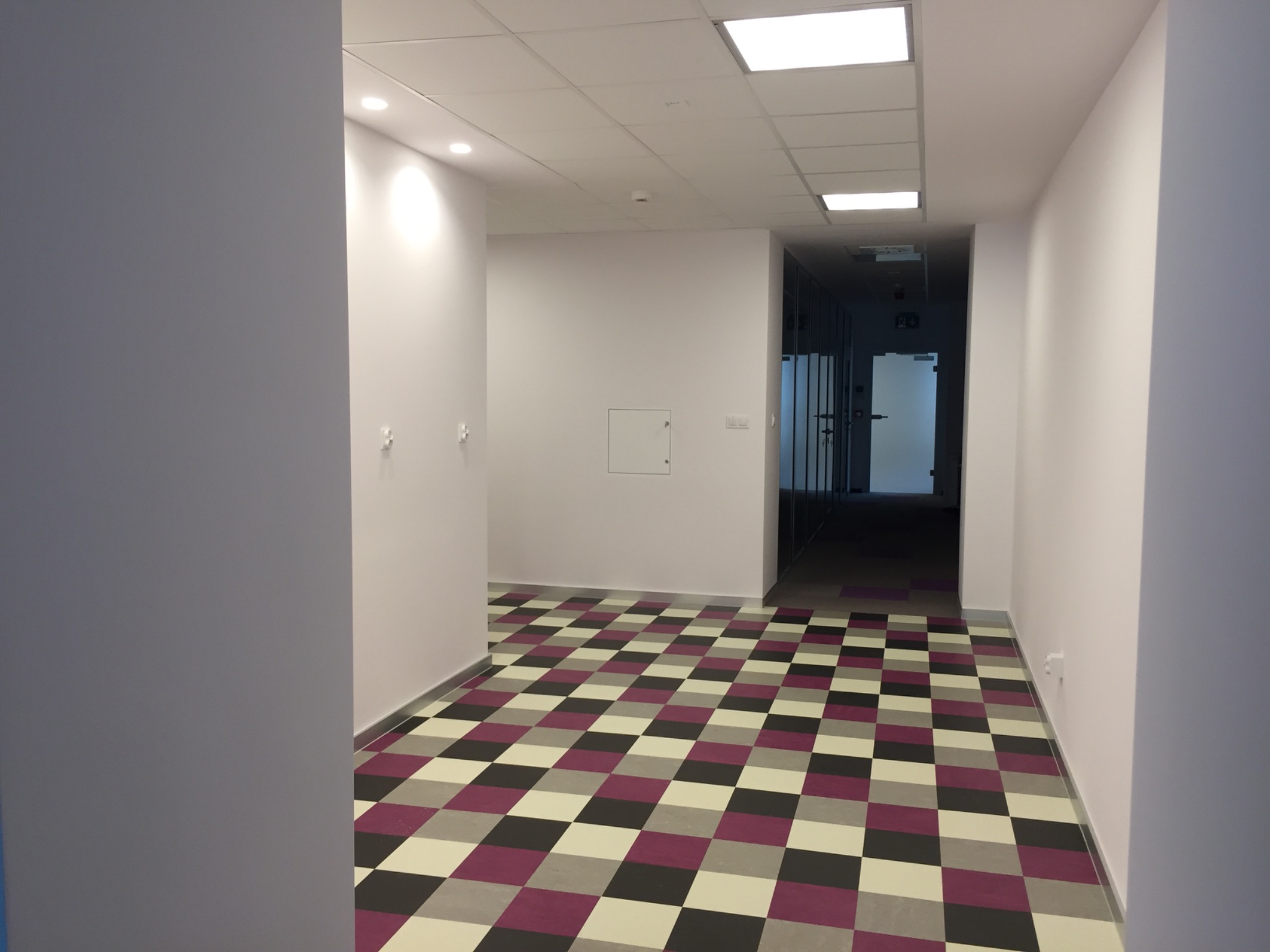 Korytarz biurowy z nową podłogą w szachownicę (czarny, szary, fioletowy), świeżo pomalowane białe ściany i oświetlenie sufitowe.